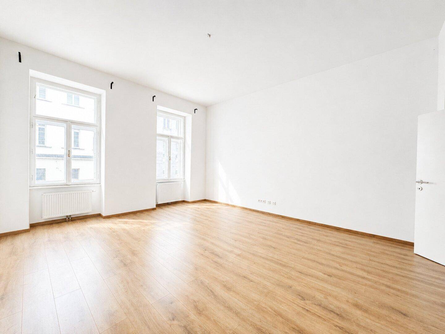 Heller, leerer Wohnraum mit Laminatboden und zwei großen Fenstern.