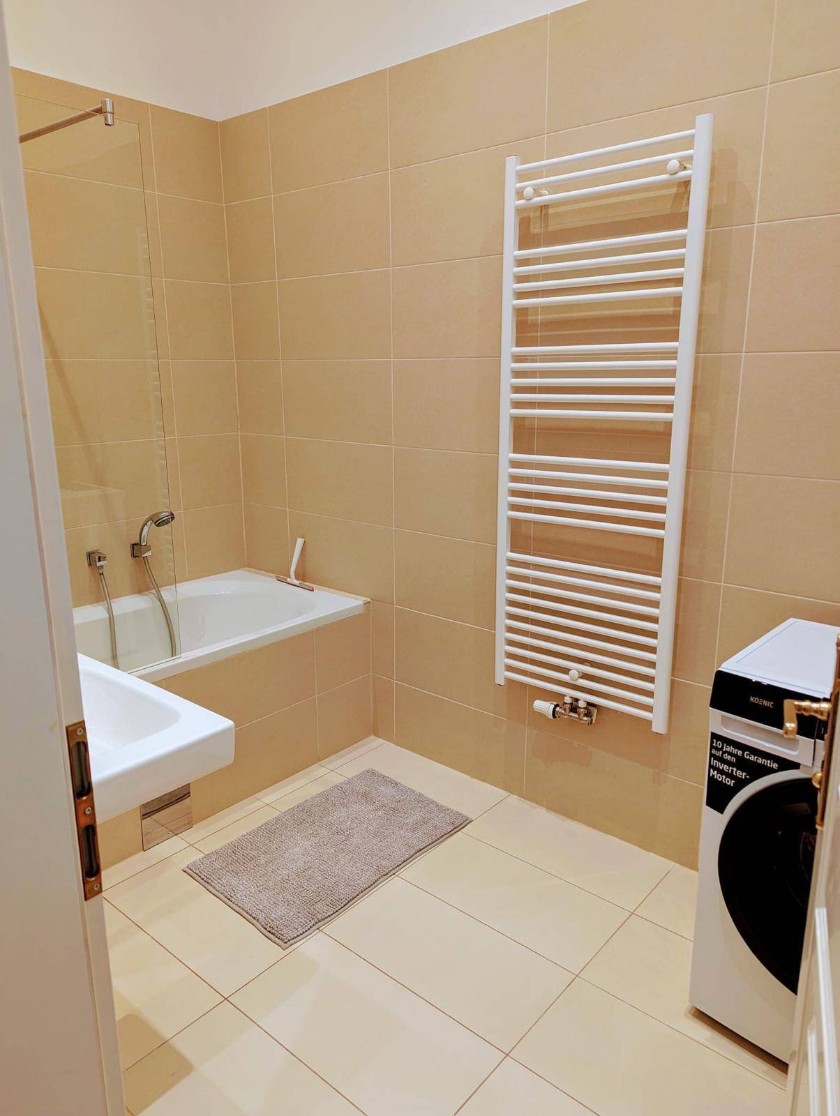 Badezimmer mit beigefarbenen Fliesen, Badewanne, Waschbecken und Handtuchheizkörper.