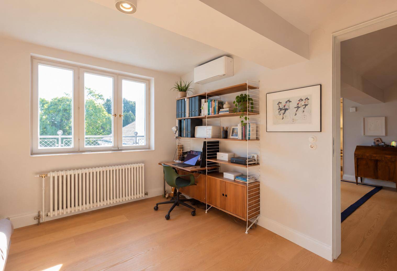 Helles Arbeitszimmer mit Holzfußboden, großem Fenster und einem modernen Regalsystem.