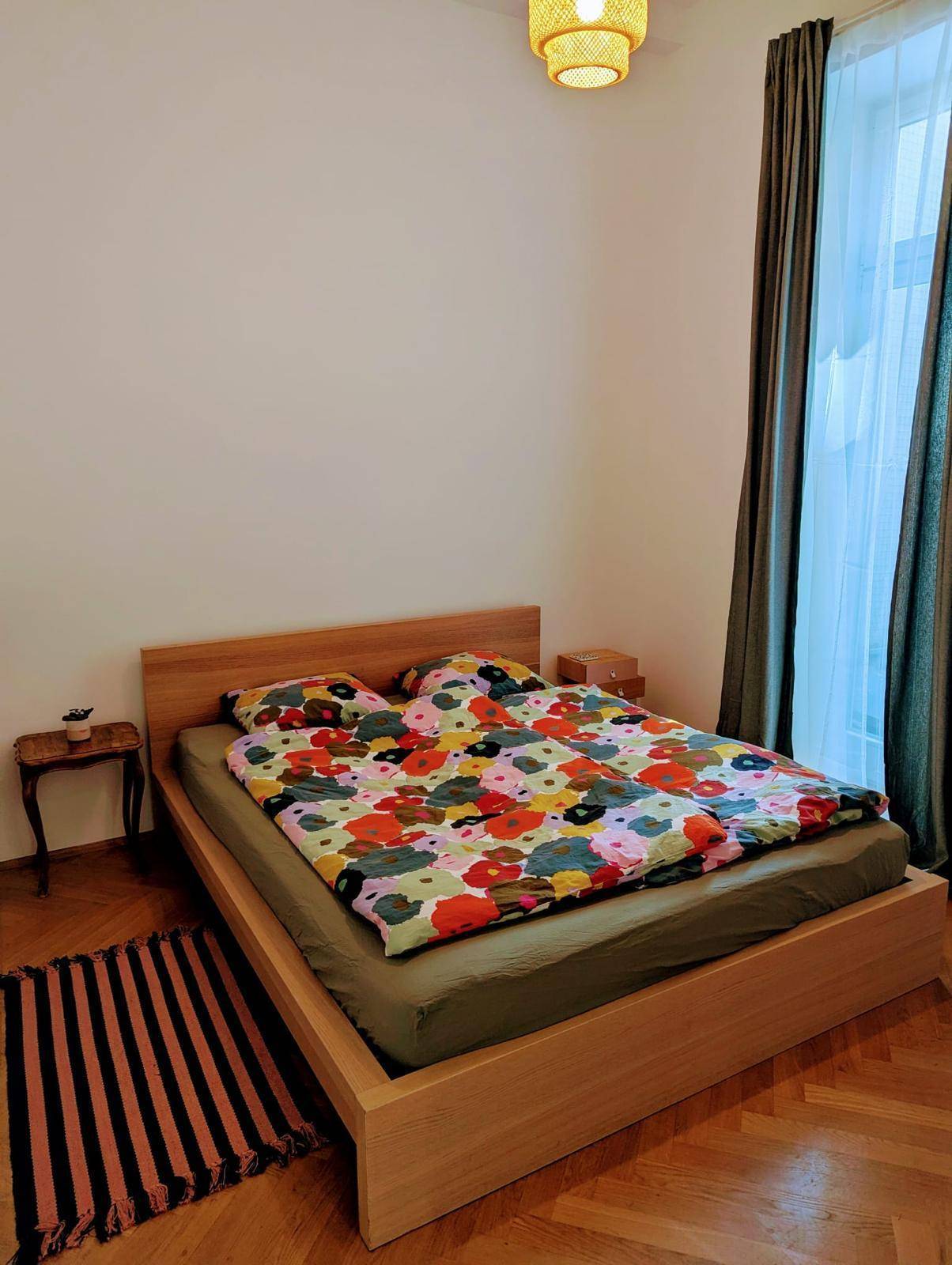 Schlafzimmer mit Holzbett, farbenfroher Bettwäsche und Fischgrätparkett, durch ein großes Fenster hell.