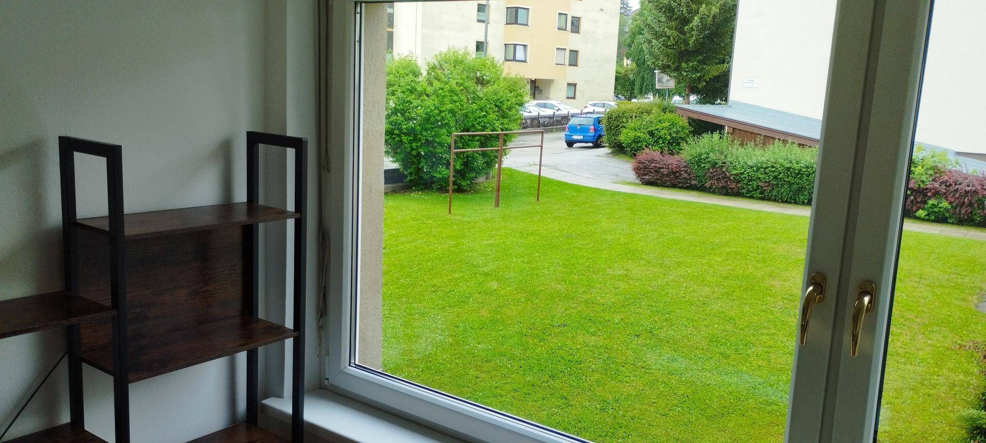 Blick aus dem Fenster auf eine grüne Rasenfläche und Bäume, mit einem Gebäude im Hintergrund.