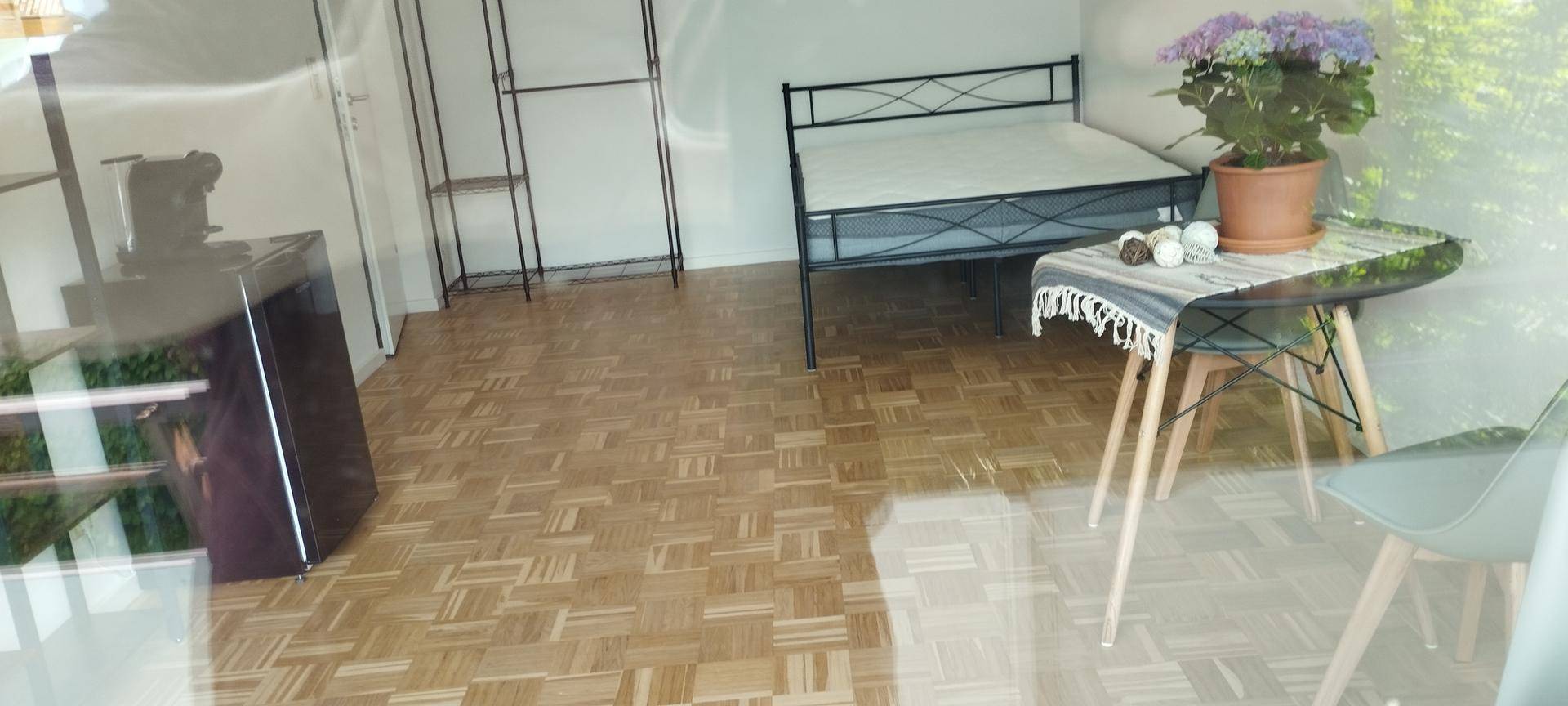 Heller Wohnraum mit Parkettboden, Bett und kleinem Kühlschrank.