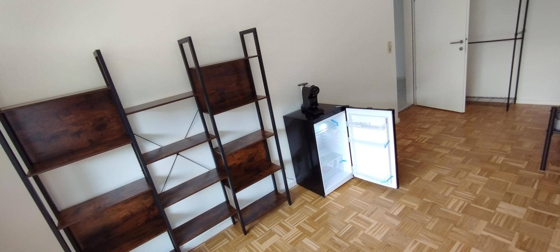 Heller Wohnraum mit Parkettboden, Regalen und einem kleinen Kühlschrank.