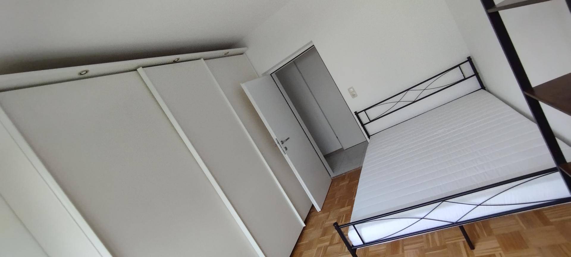 Schlafzimmer mit Bettgestell, Matratze und Einbauschränken, Parkettboden und Zugang zu einem weiteren Raum.