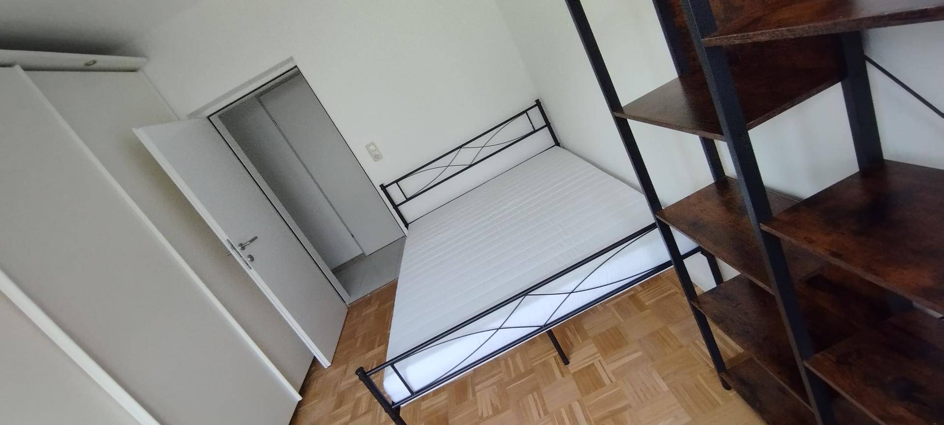 Schlafzimmer mit Bettgestell, Matratze und Regal, Parkettboden und Zugang zu einem weiteren Raum.