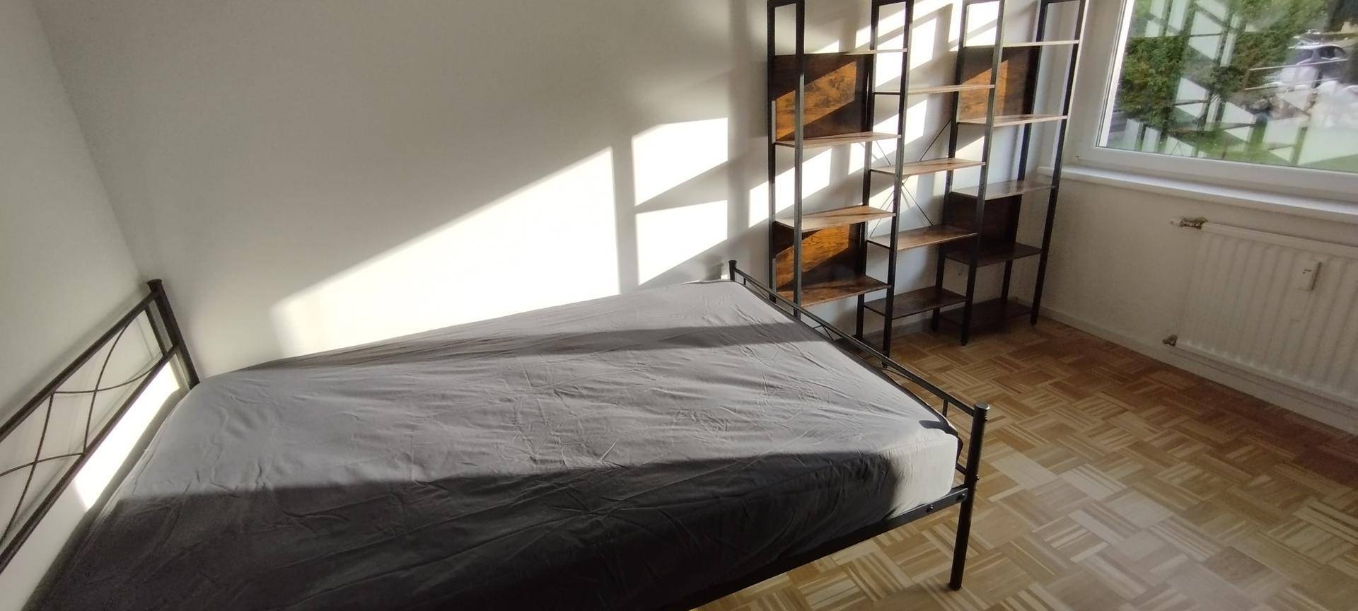 Schlafzimmer mit Holzboden, einem einfachen Bett und einem Regal, beleuchtet durch das Fenster.