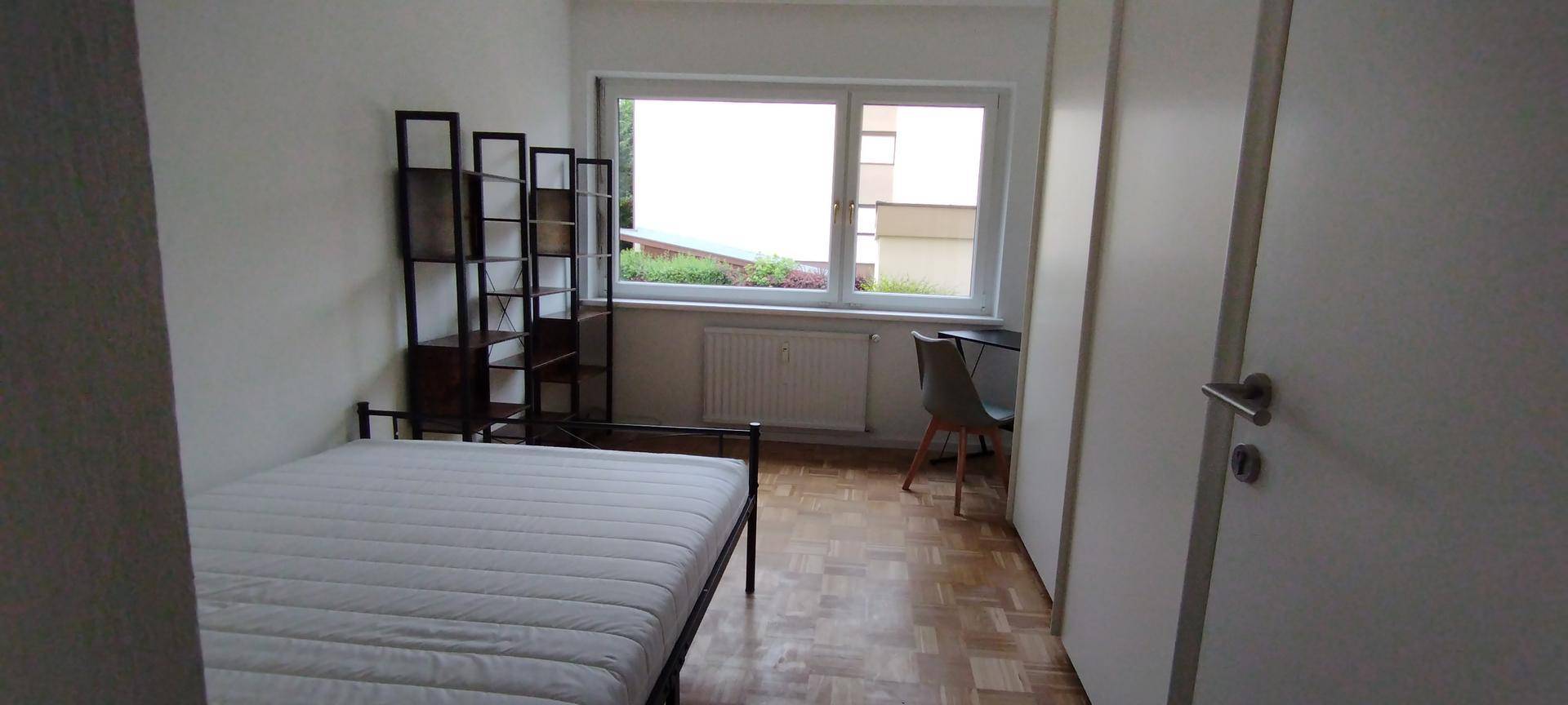 Helles Schlafzimmer mit Bett, Regal, Schreibtisch und Stuhl, mit Blick aus dem Fenster.