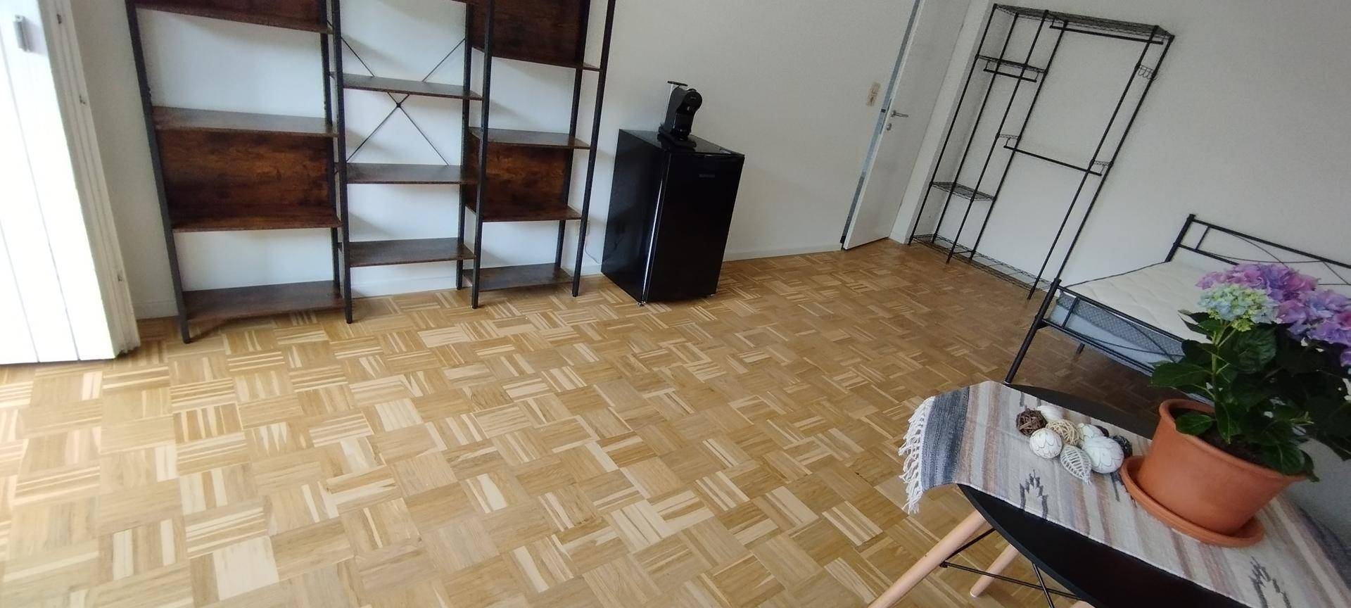 Heller Wohnraum mit Parkettboden, Regalen und kleinem Kühlschrank.