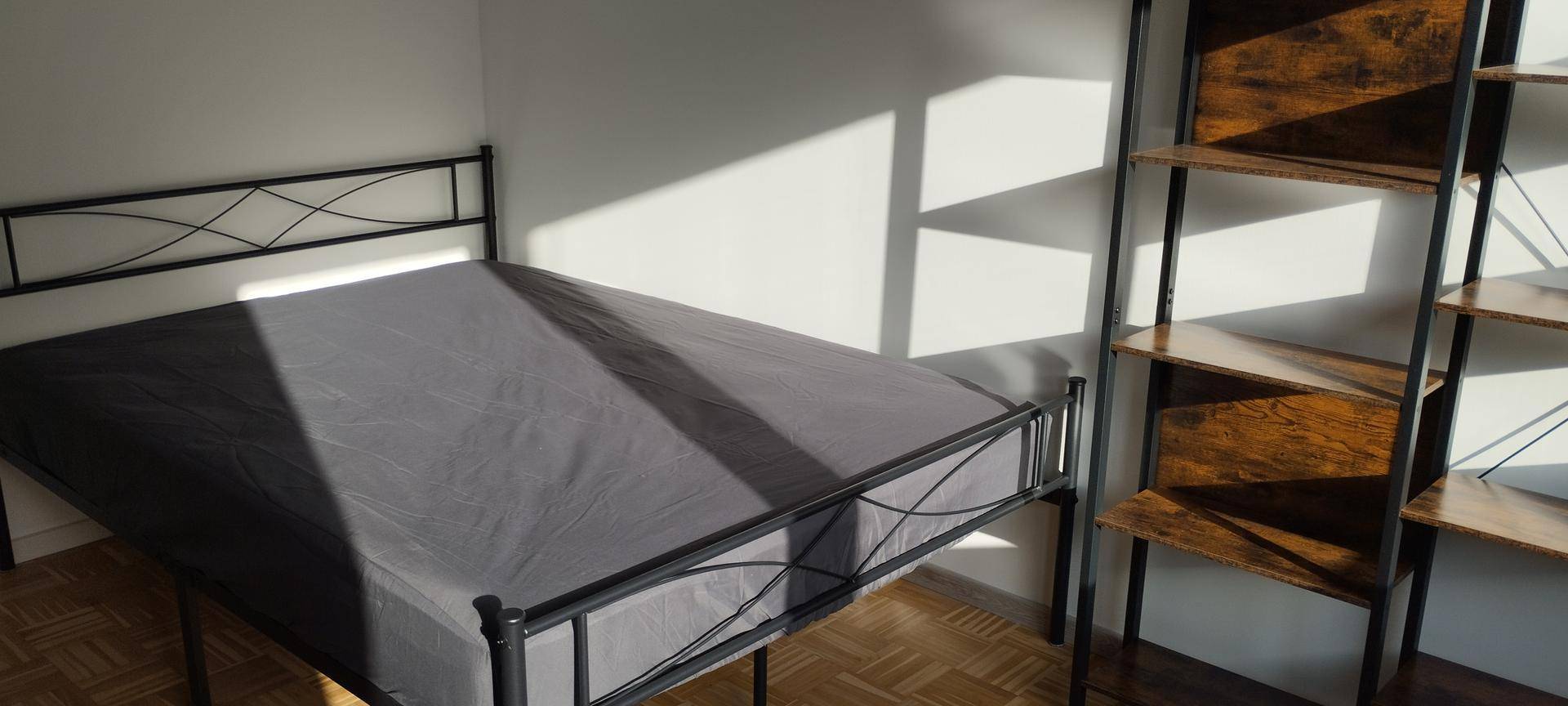 Helles Schlafzimmer mit Parkettboden, einem Bettgestell und einem hohen Regal, durchflutet von Sonnenlicht.