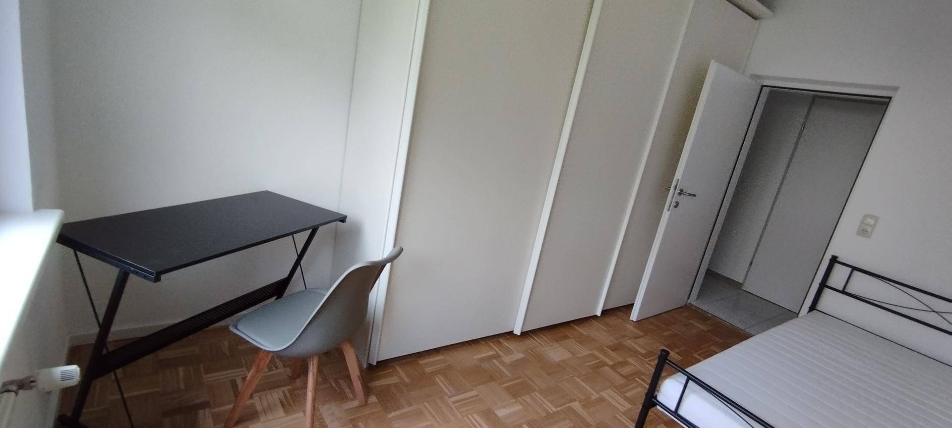 Schlafzimmer mit Schreibtisch, Stuhl und Einbauschränken, Parkettboden und Zugang zu einem weiteren Raum.