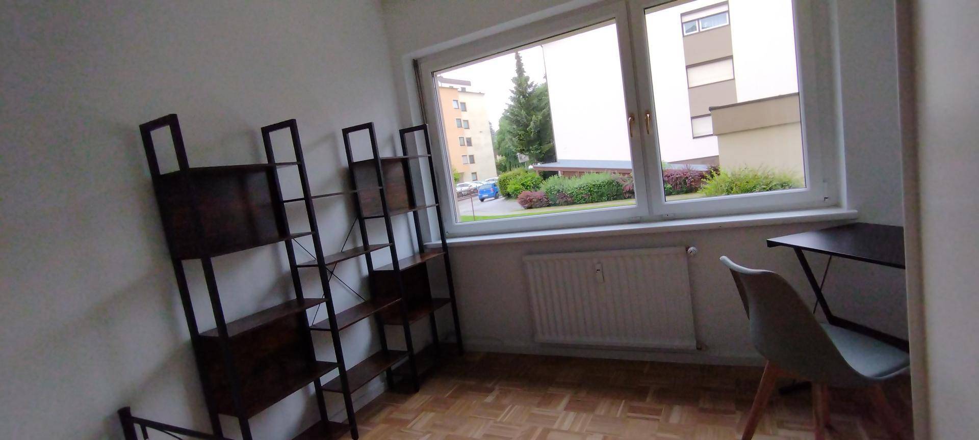 Schlafzimmer mit Regal, Schreibtisch und Stuhl, bietet einen Ausblick auf Gebäude und Grünflächen.