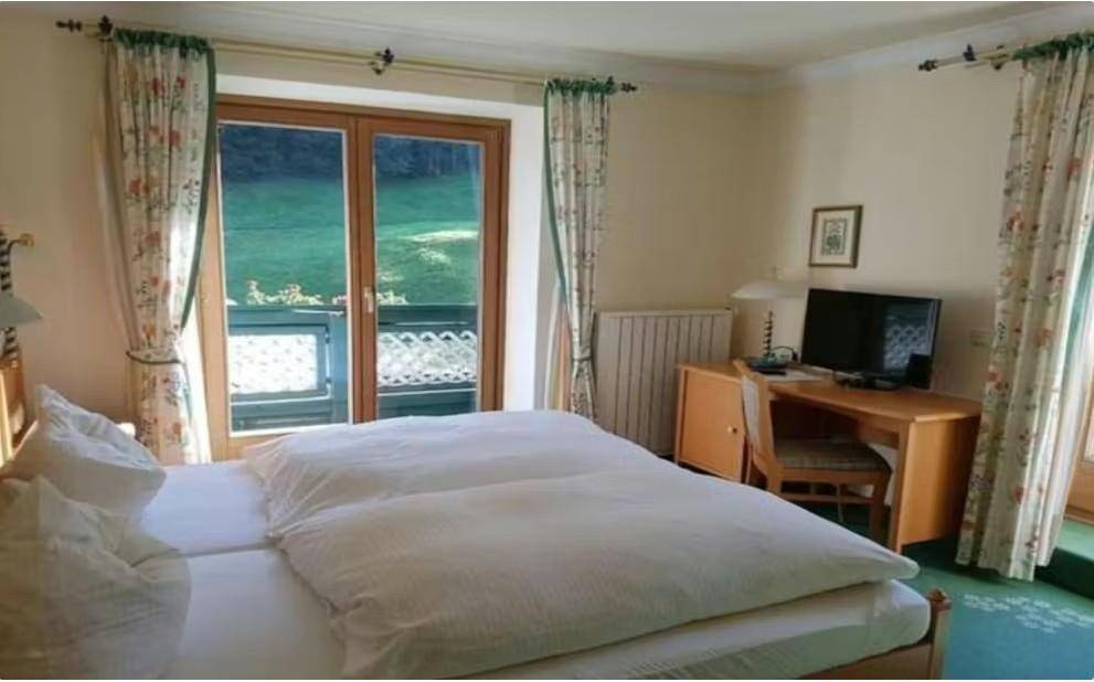 Helles Schlafzimmer mit Doppelbett, Schreibtisch und Zugang zu einem Balkon mit Blick ins Grüne.