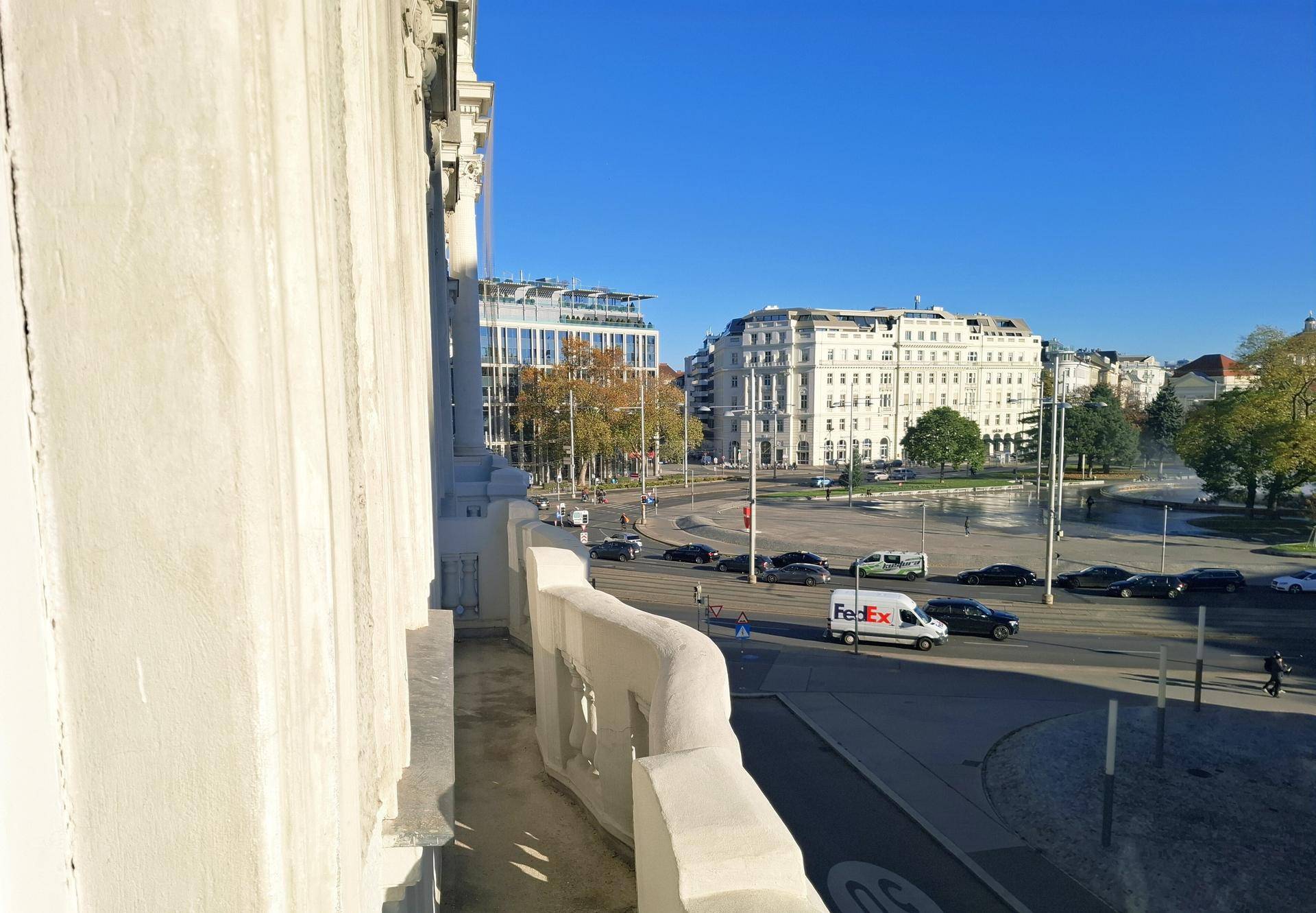 Weiter Blick von einem Balkon über eine städtische Kreuzung mit Verkehr und umliegenden Gebäuden an einem sonnigen Tag.