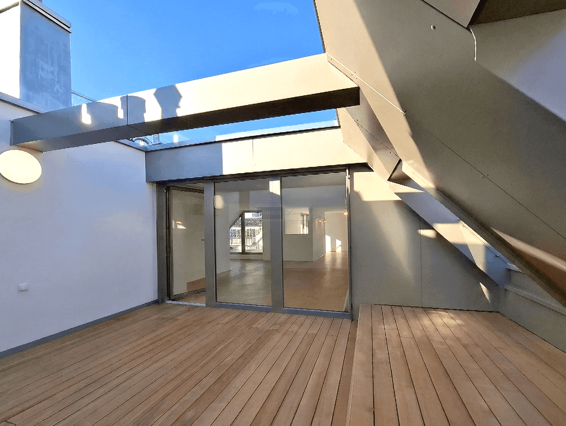 Weitläufige Dachterrasse mit Holzdielen und großzügigen Glasfronten zum Wohnraum, unter blauem Himmel.