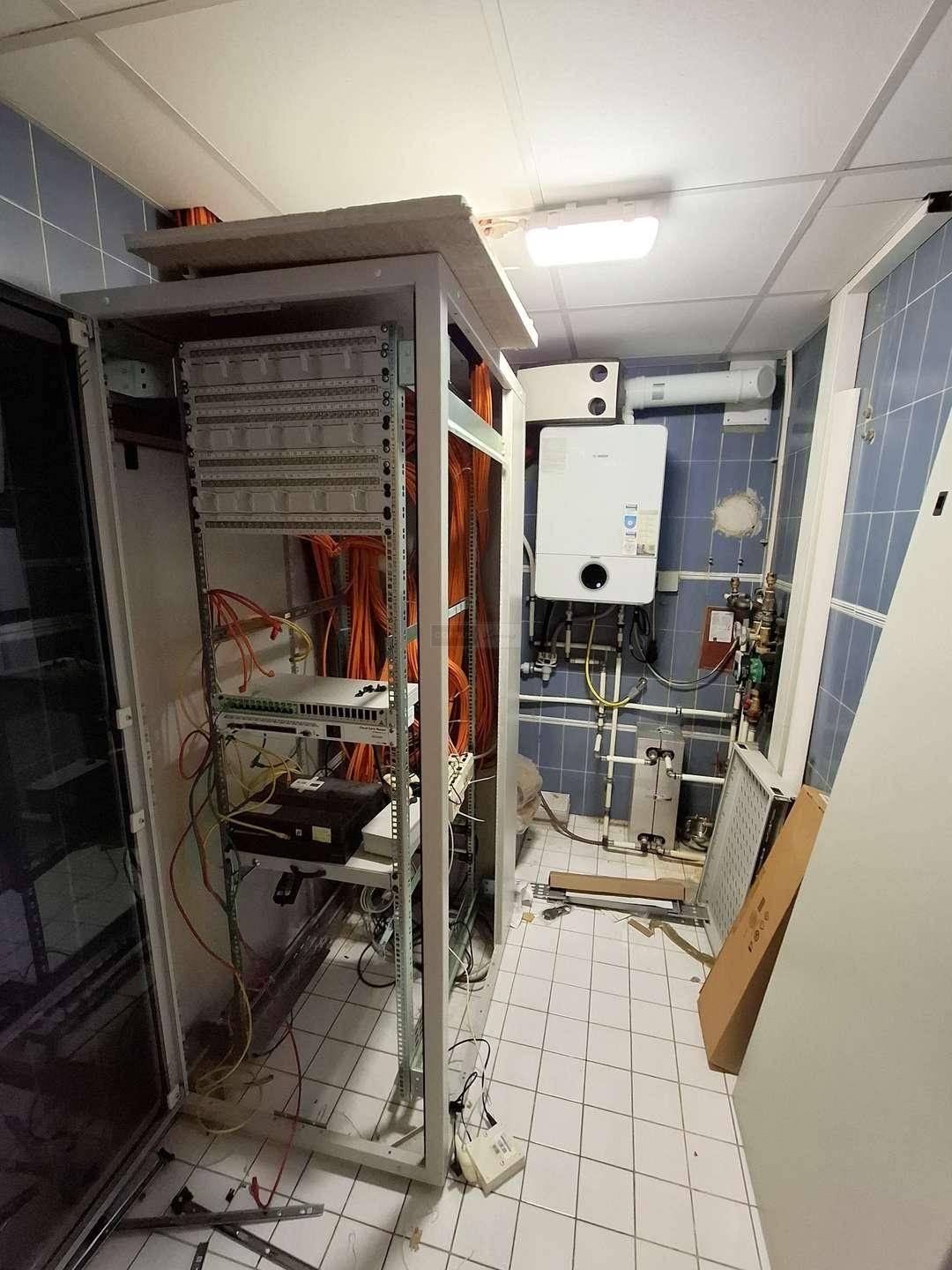 Technikraum mit Serverrack, sichtbaren Kabeln, Warmwasserbereiter und gefliesten Wänden und Böden.