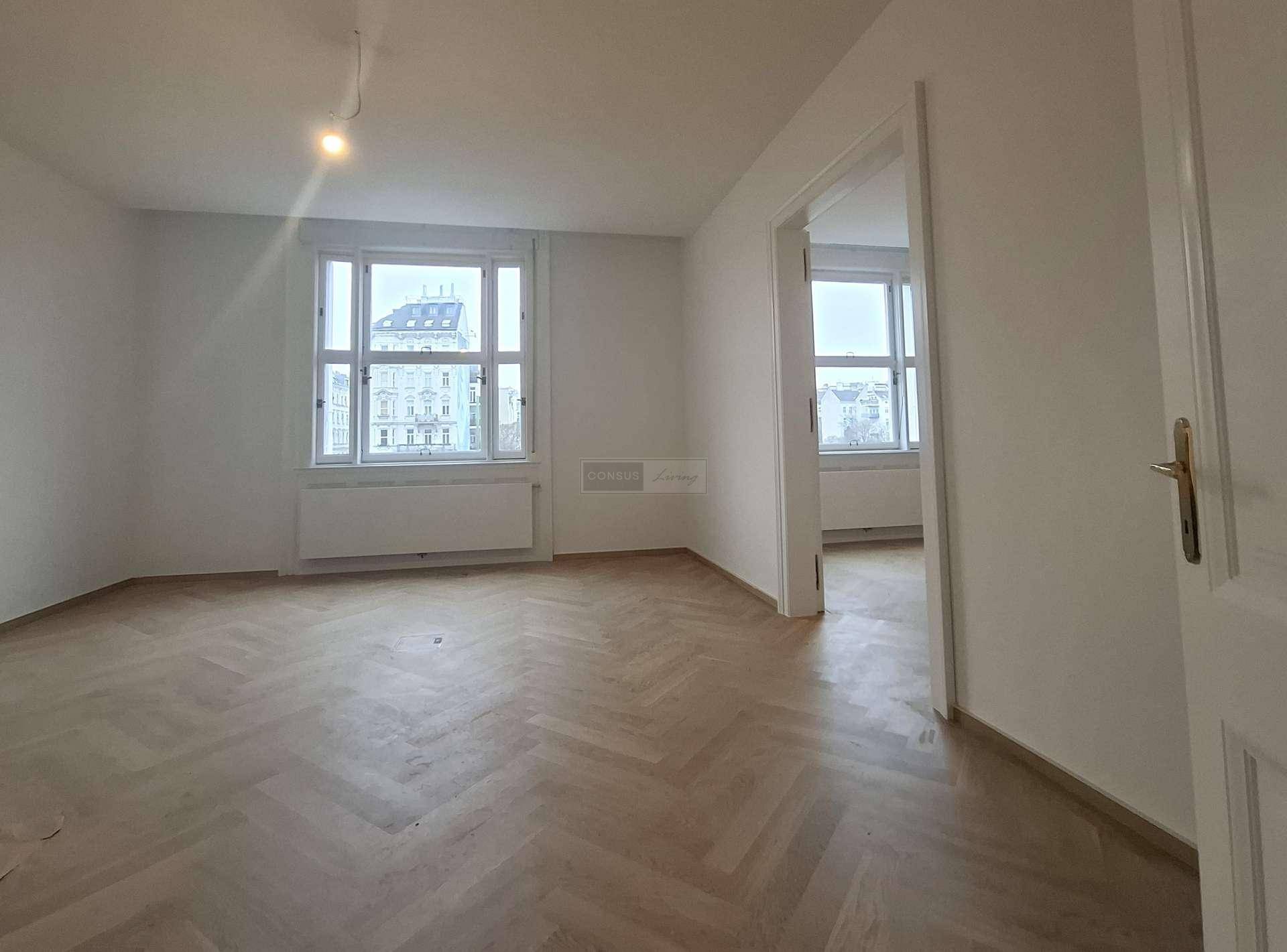 Helles, leeres Zimmer mit Fischgrätparkett und Fenstern, die einen Blick auf die umliegenden Gebäude ermöglichen.