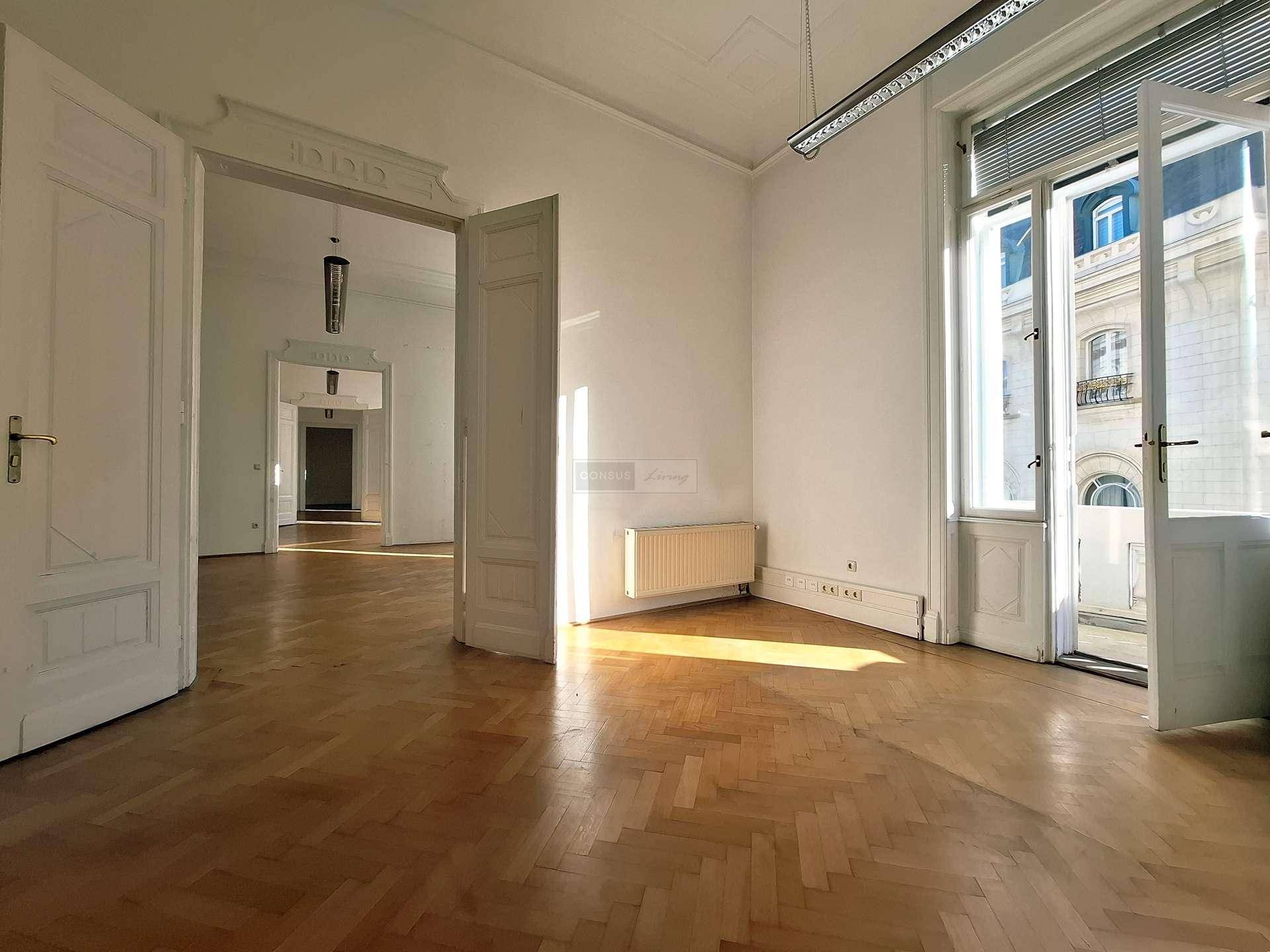 Großer, heller Raum mit Fischgrätparkett, hohen Decken und einer geöffneten Balkontür, die Tageslicht hereinlässt.
