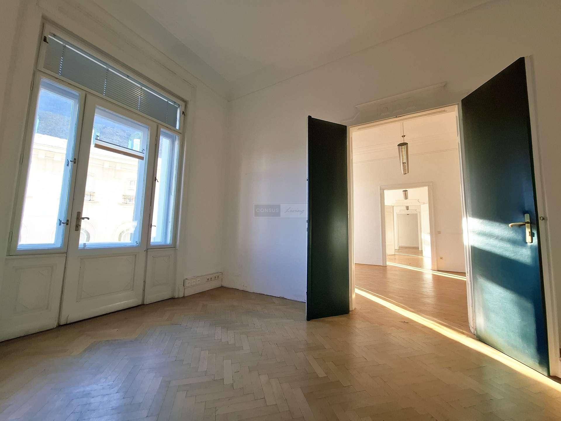Leerer Raum mit Fischgrätparkett, hohen Fenstern und Blick in einen weiteren, sonnendurchfluteten Raum.