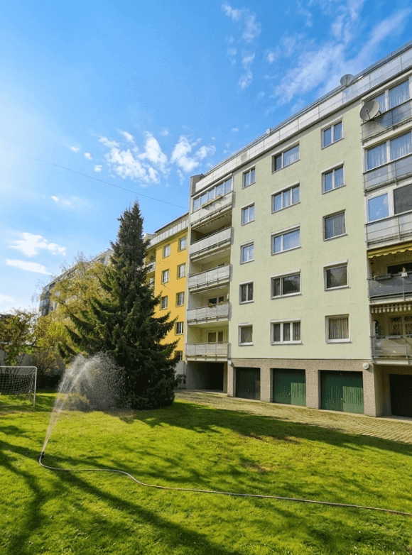 Mehrfamilienhaus mit grüner Rasenfläche, Bäumen und Garagen im Erdgeschoss.
