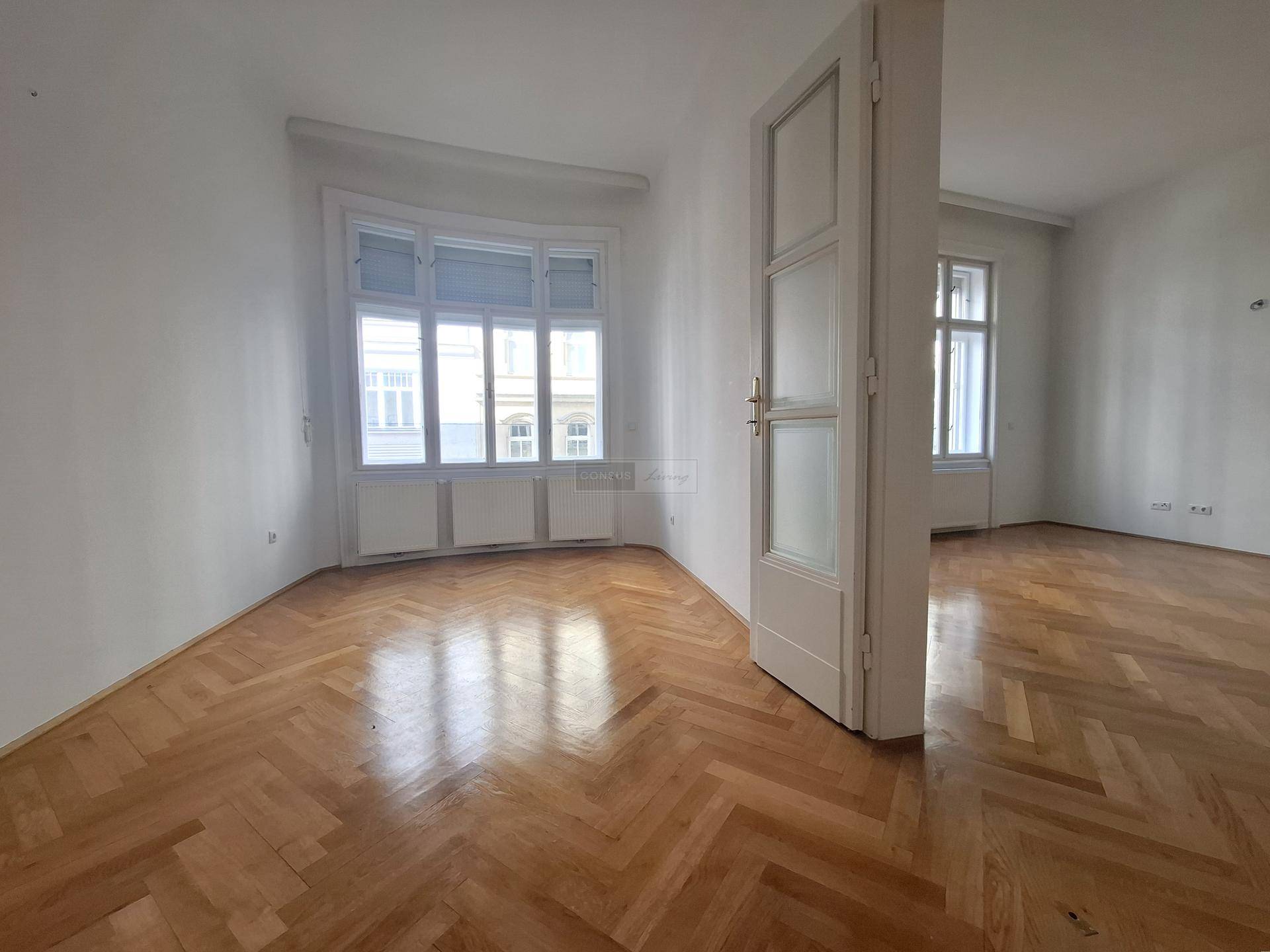 Geräumiges Zimmer mit Fischgrätparkett und großen Fenstern, das viel Tageslicht hereinlässt.