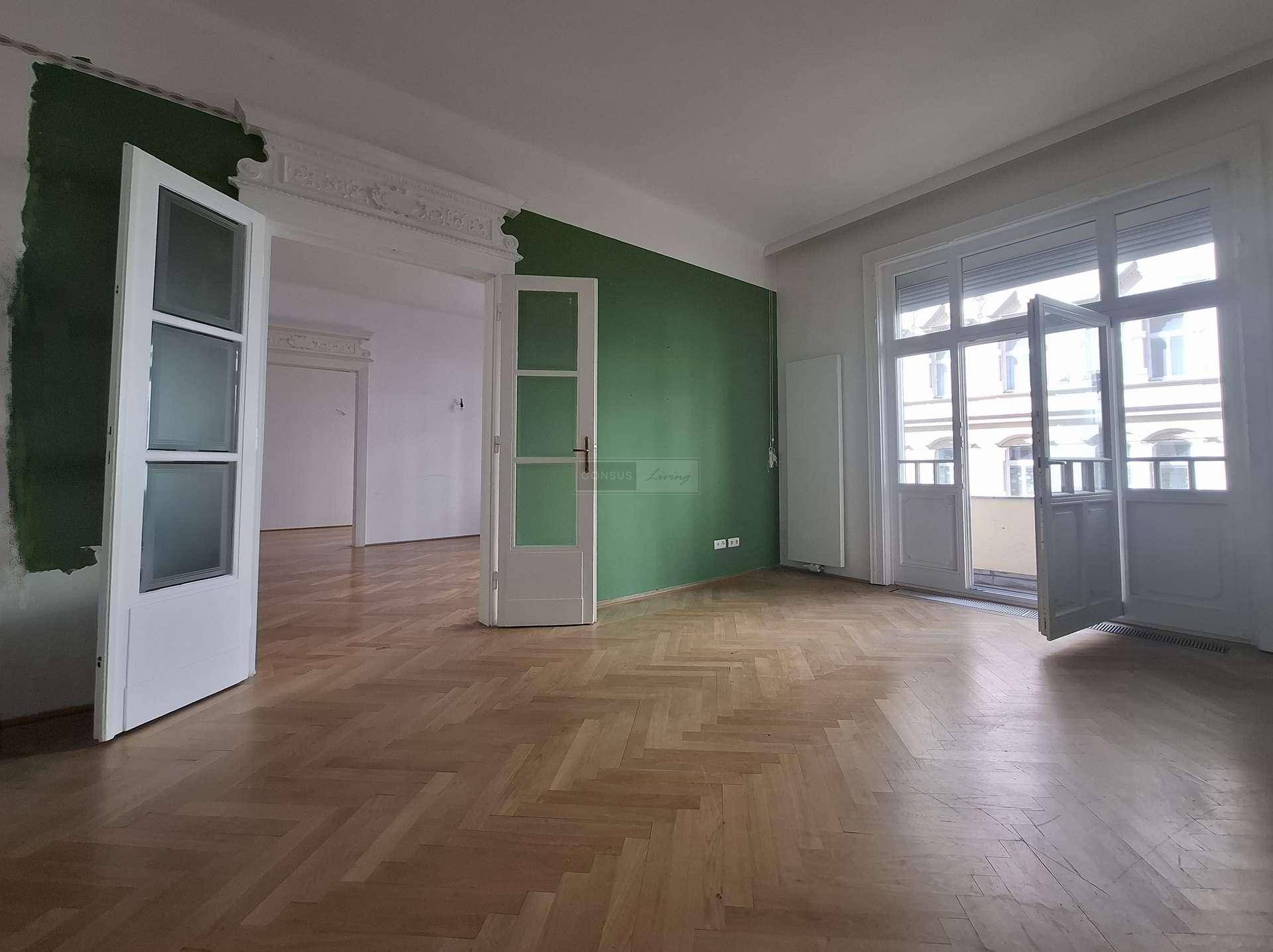 Geräumiges Wohnzimmer mit Fischgrätparkett, grüner Akzentwand und Zugang zum Balkon.