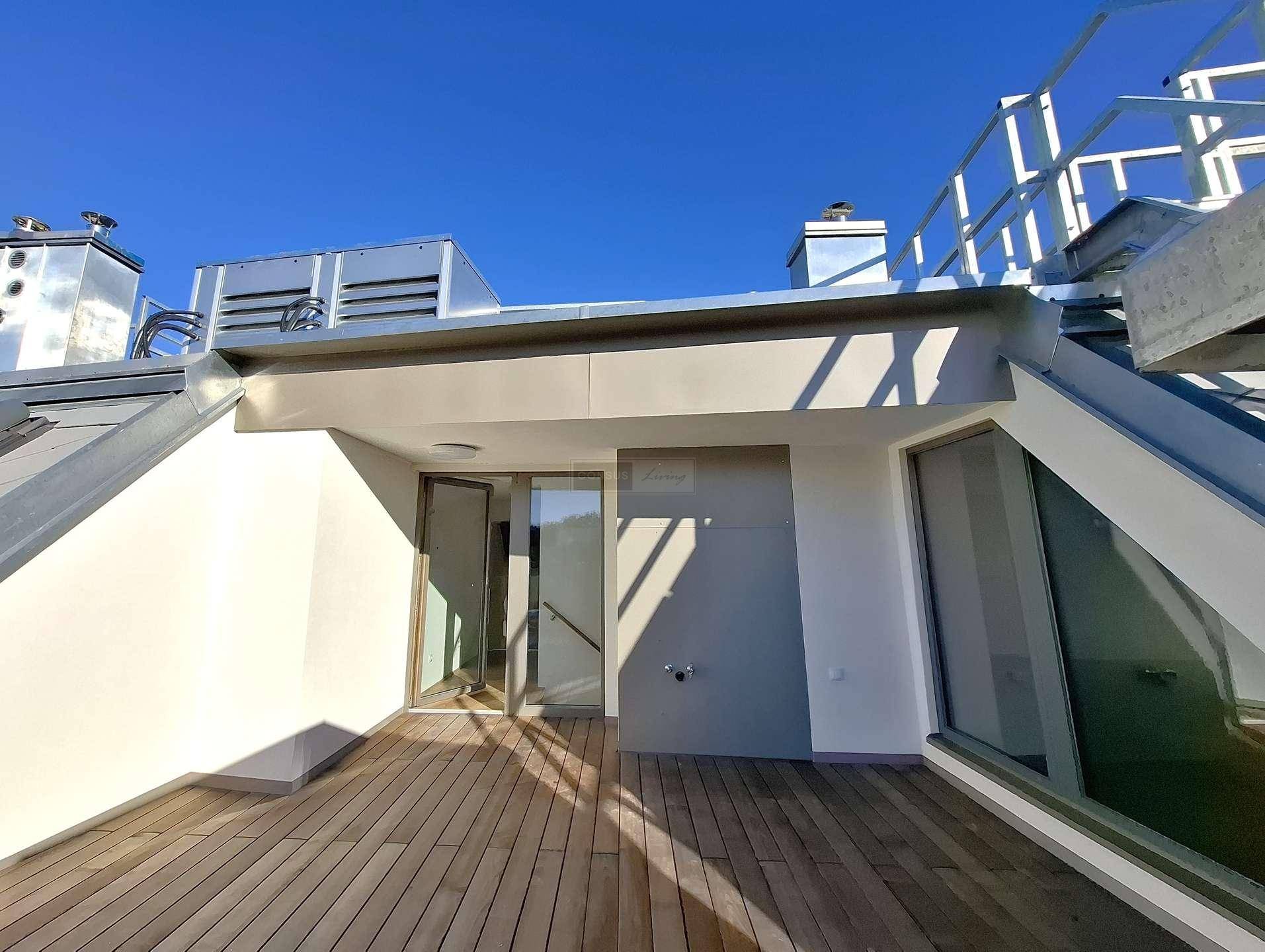 Zugang zur Dachterrasse mit Holzboden und Blick auf die umliegenden Gebäude unter blauem Himmel.