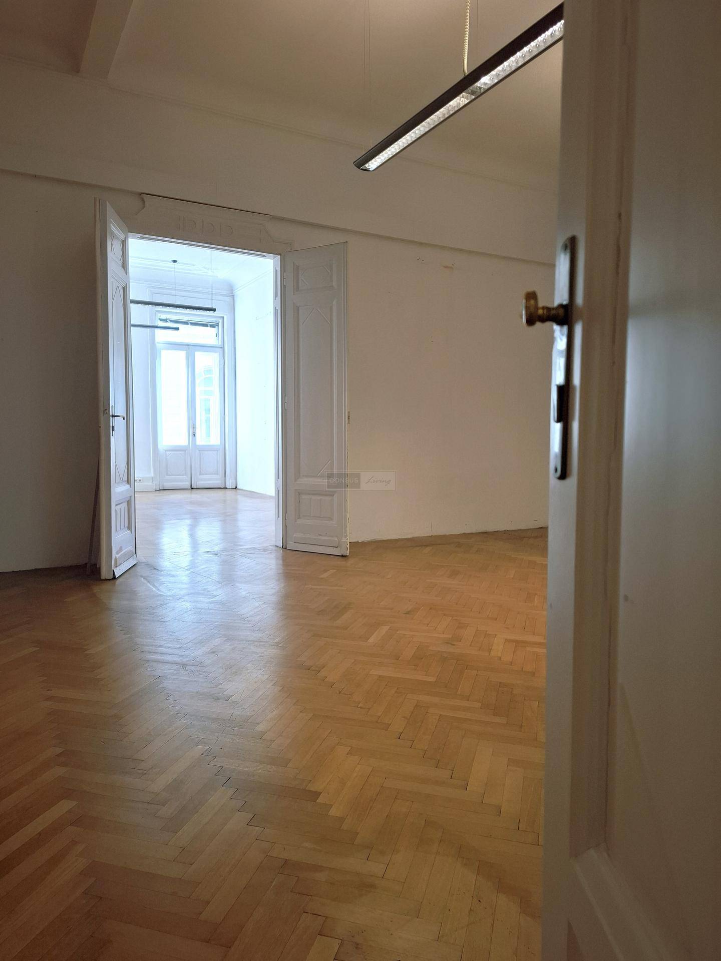 Blick durch eine offene Tür in einen weiteren leeren Raum mit klassischem Fischgrätparkett und hohen Decken.