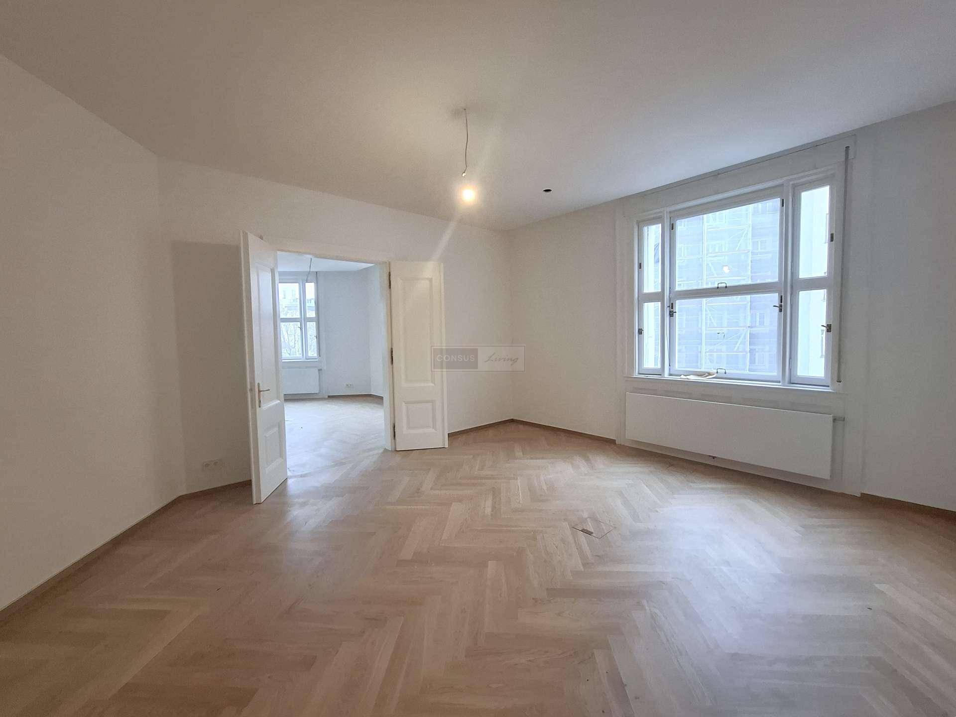 Geräumiges Zimmer mit Fischgrätparkett und großen Fenstern, das viel Tageslicht empfängt.