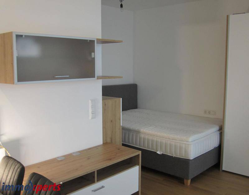 Schlafbereich mit Bett, Schrank und Schreibtisch in einem hellen Raum, praktisch eingerichtet.