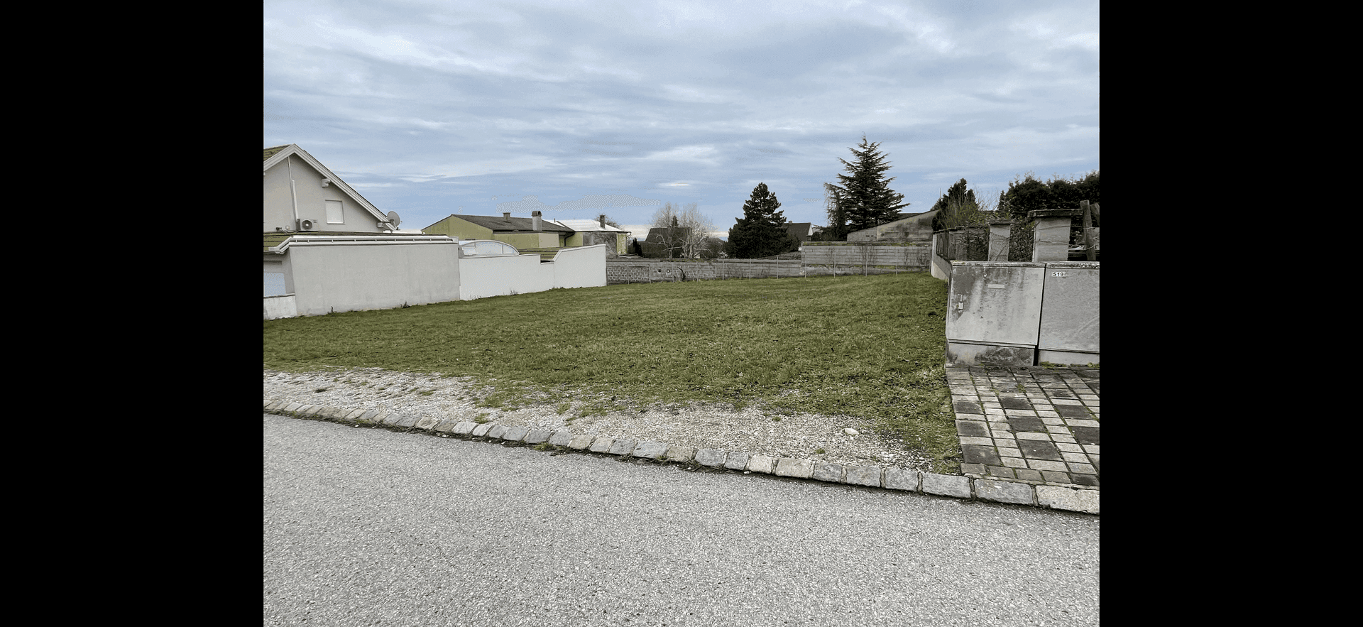 Unbebautes Grundstück mit Rasenfläche und angrenzenden Gebäuden unter bewölktem Himmel.