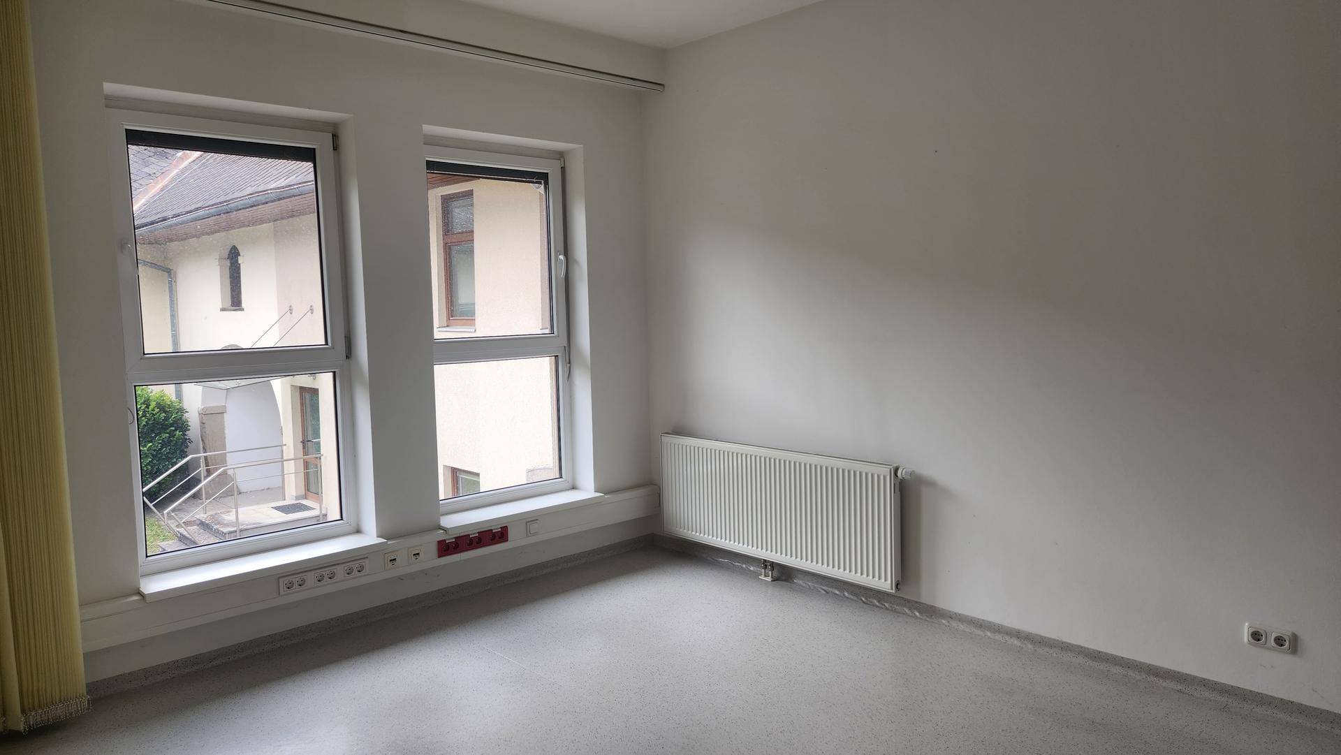Einfacher Büroraum mit zwei Fenstern, die Tageslicht hereinlassen, und einem Heizkörper.
