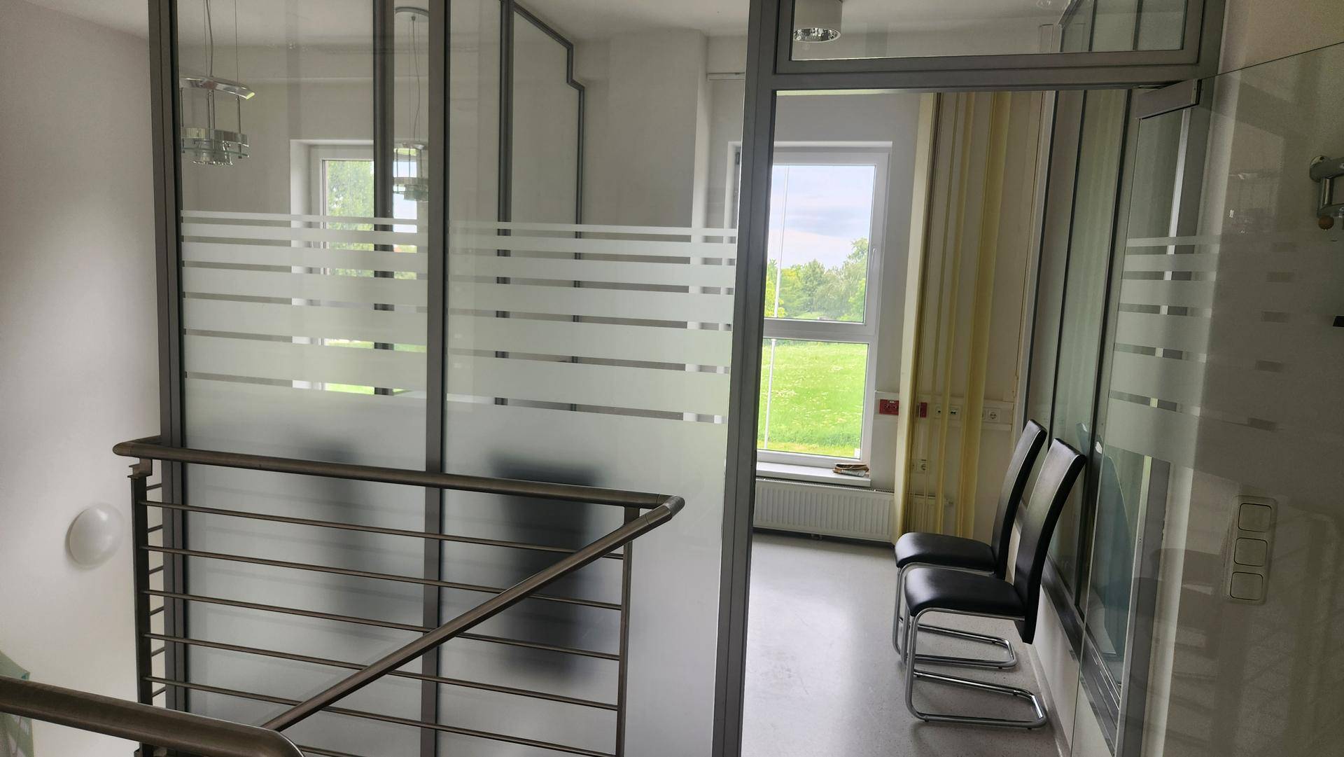 Treppenbereich mit Metallgeländer und Blick in einen hellen Büroraum mit großen Fenstern.