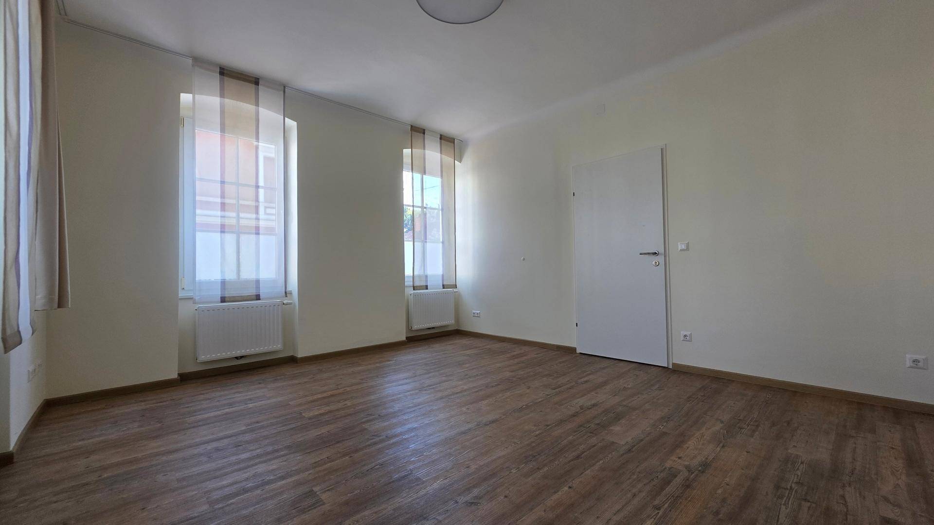 Geräumiger Wohnbereich mit Laminatboden und zwei großen Fenstern, die viel Tageslicht hereinlassen.