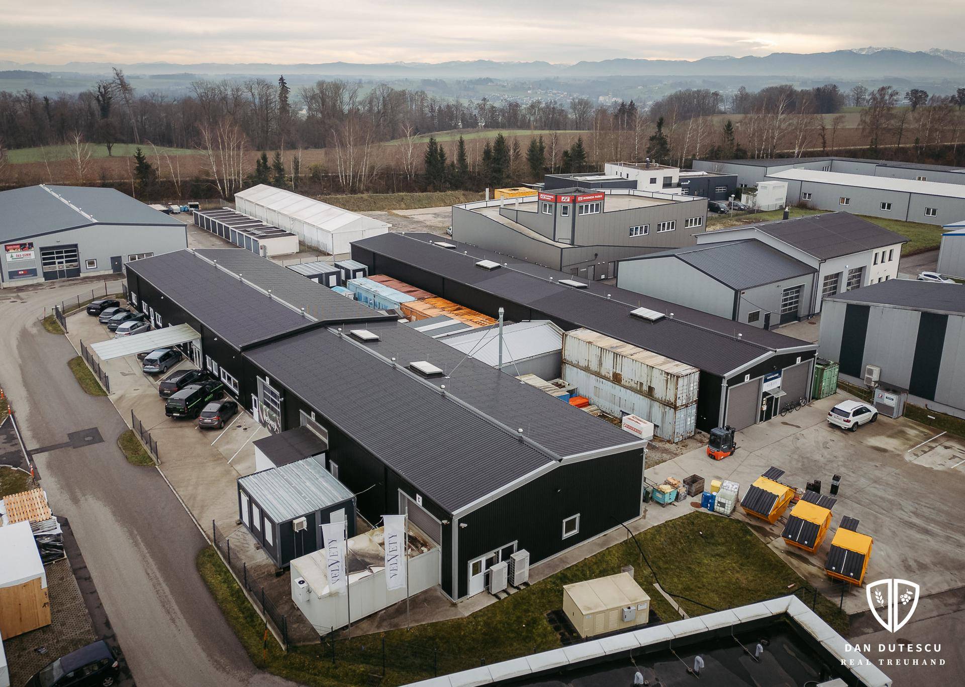 Luftaufnahme eines Gewerbekomplexes mit mehreren Hallen, Parkplätzen und umliegender Landschaft unter bewölktem Himmel.