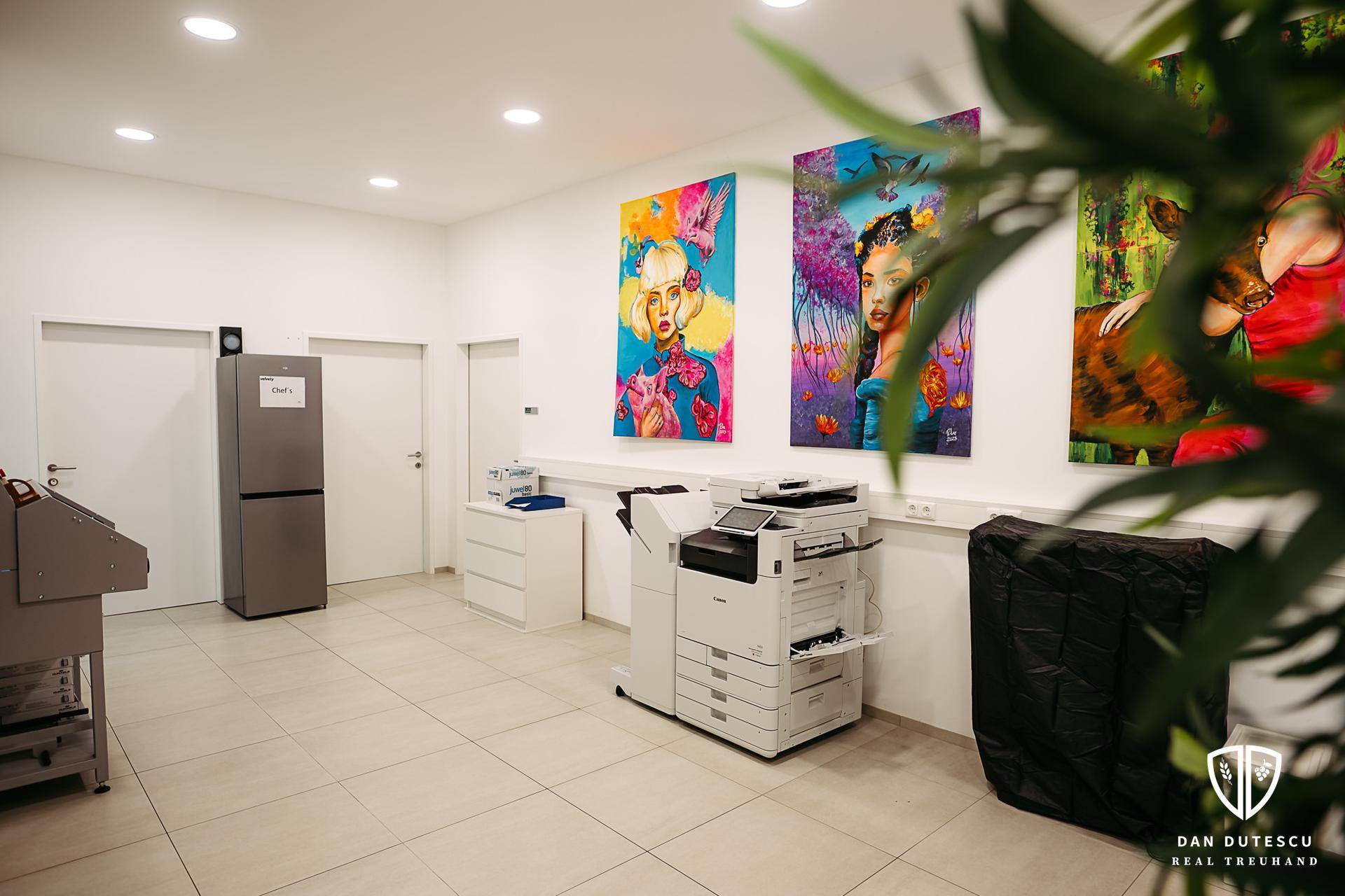 Heller Bürobereich mit Drucker, Kühlschrank und farbenfrohen Kunstwerken an den Wänden.