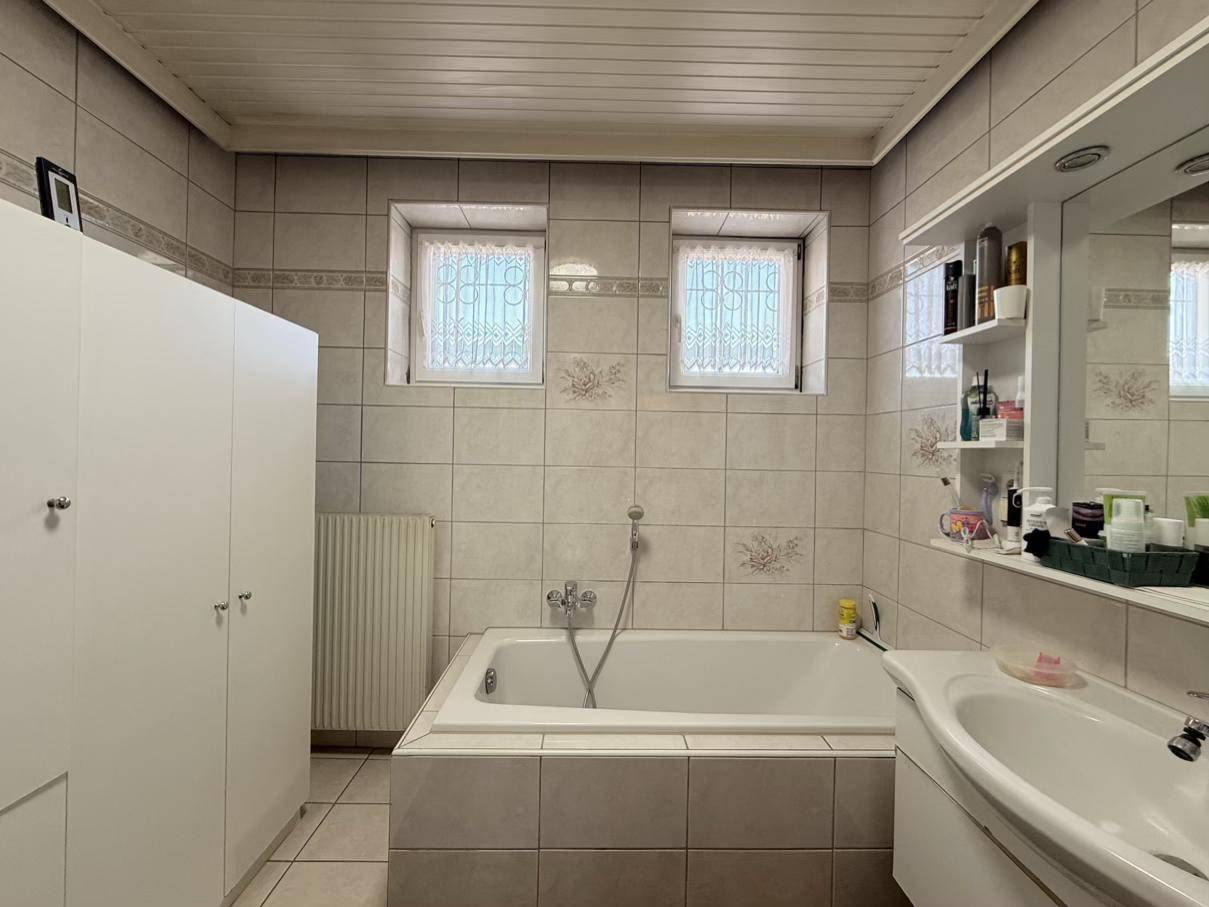 Gefliestes Badezimmer mit Badewanne, Waschbecken und Spiegelschrank, beleuchtet durch zwei Fenster.