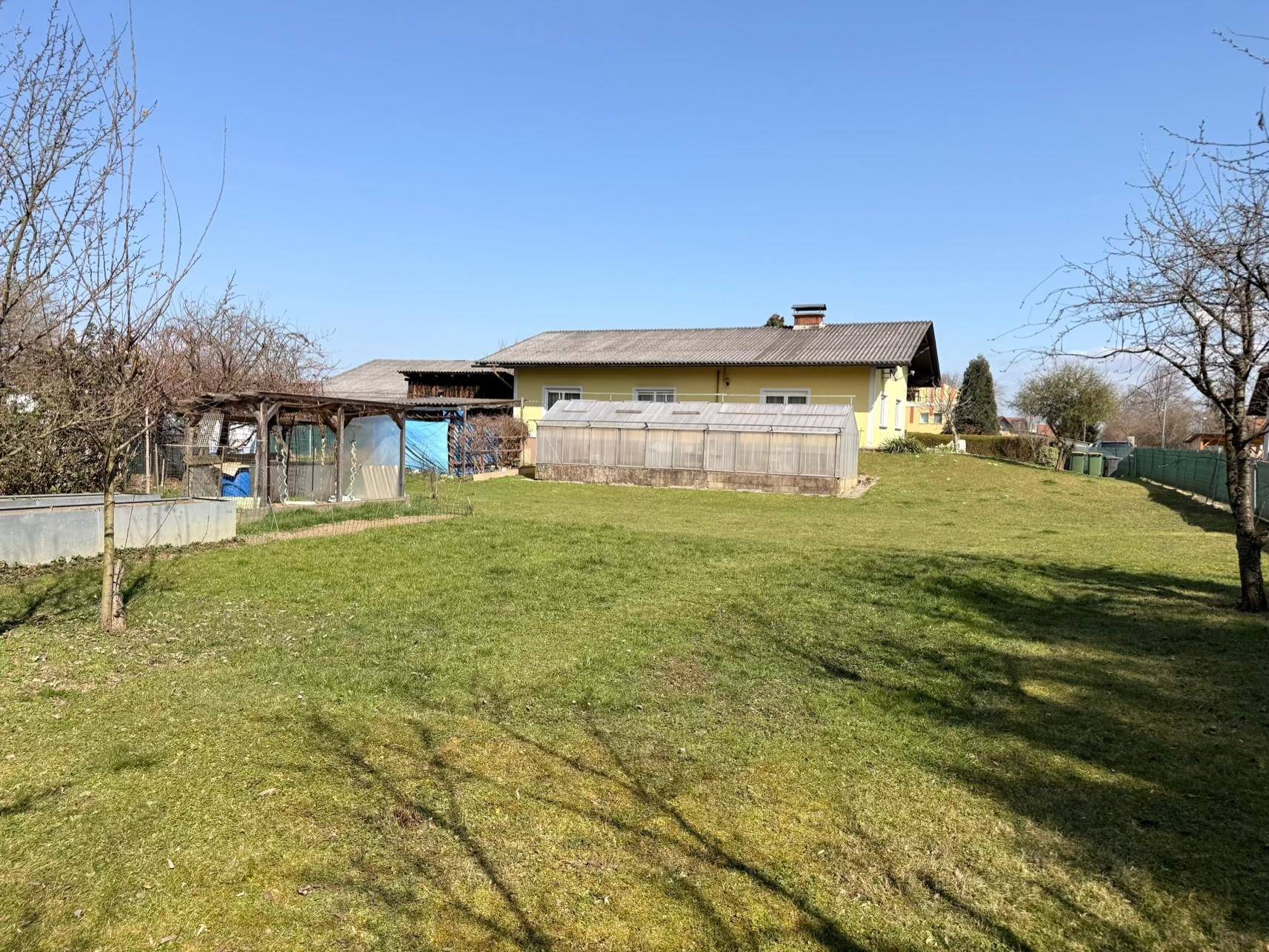 Weitläufiger Garten mit Rasenfläche, Gewächshaus und dem gelben Haus im Hintergrund an einem sonnigen Tag.