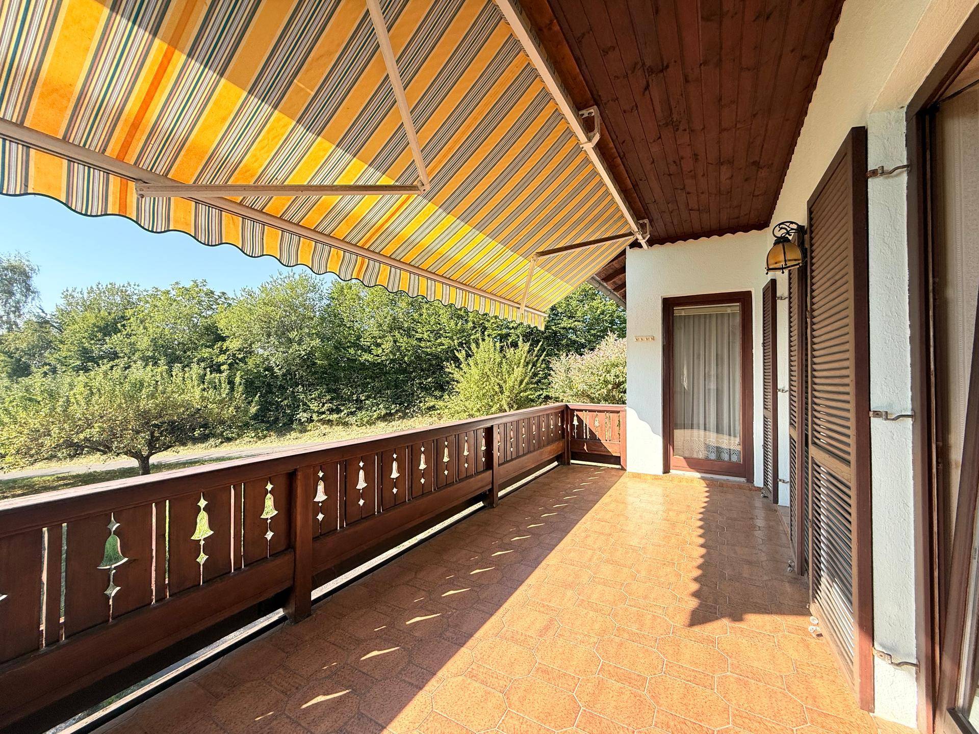 Sonniger Balkon mit Holzgeländer, Fliesenboden und gestreifter Markise, umgeben von Grün.