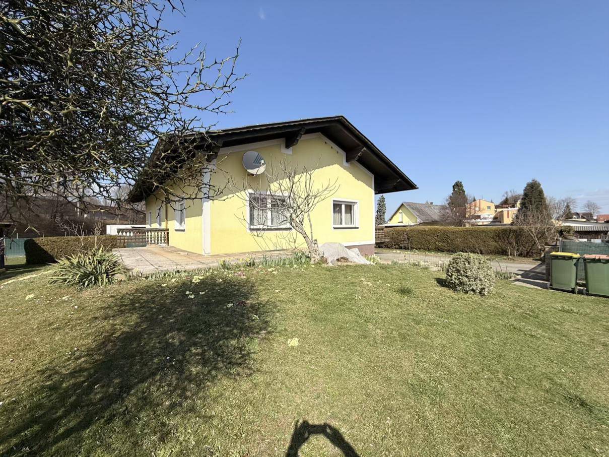 Ansicht des gelben Einfamilienhauses von der Seite mit einem grünen Rasenhügel und Mülltonnen im Vordergrund.