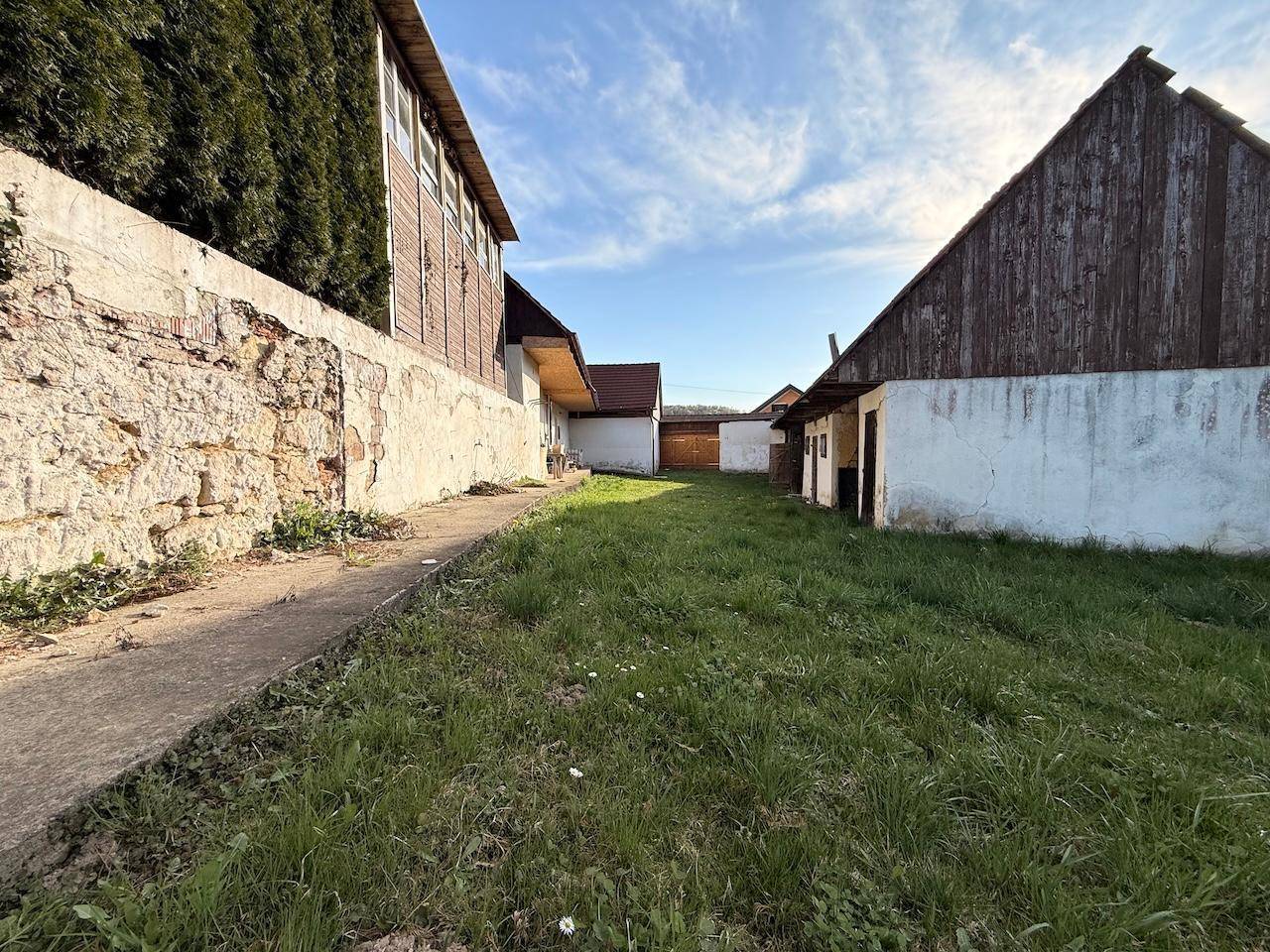 Weg zwischen ländlichen Gebäuden mit Grasflächen und einer alten Steinmauer unter blauem Himmel.