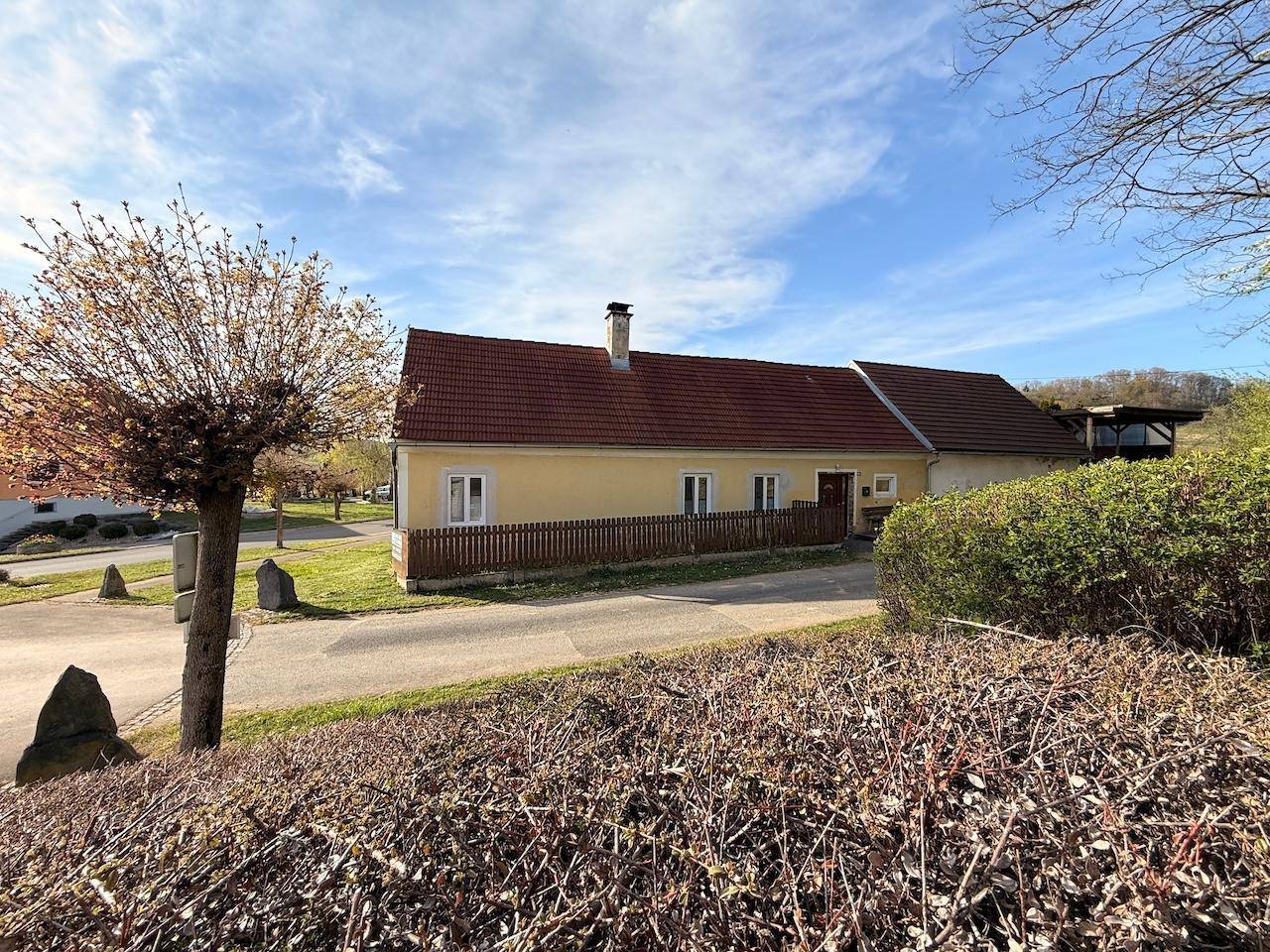 Gelbes Landhaus mit rotem Dach, umgeben von einer Hecke und Bäumen in einer ländlichen Landschaft.