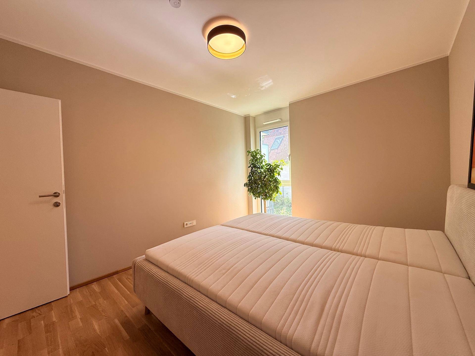 Schlafzimmer mit Doppelbett, Parkettboden und Fenster, das viel Tageslicht hereinlässt.