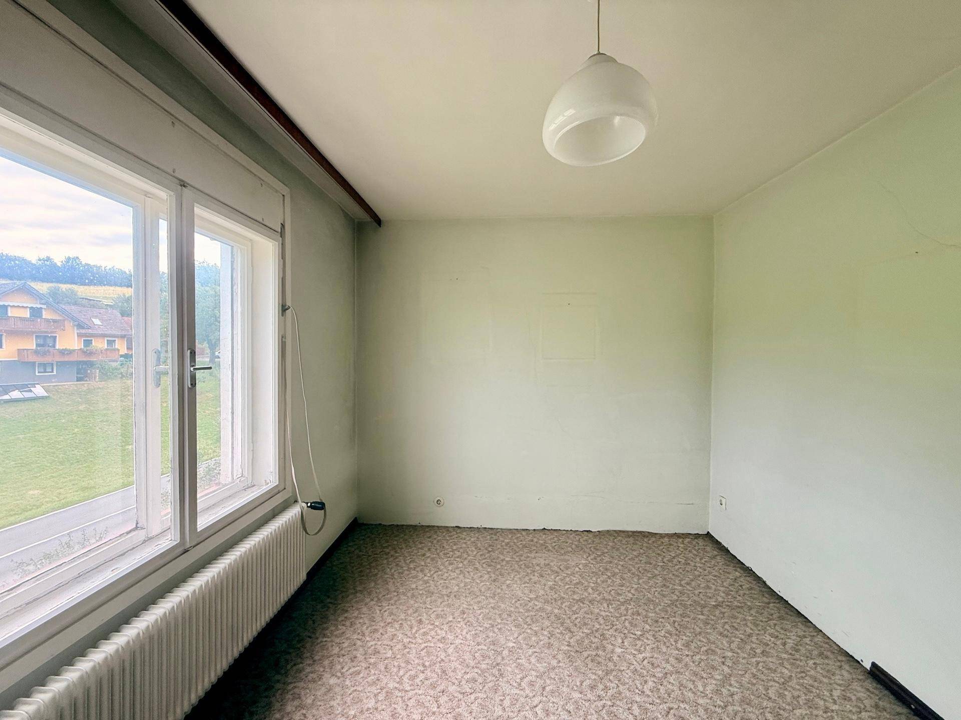 Einfaches Zimmer mit Teppichboden, großem Fenster und Heizkörper, bietet einen weiten Blick ins Grüne.