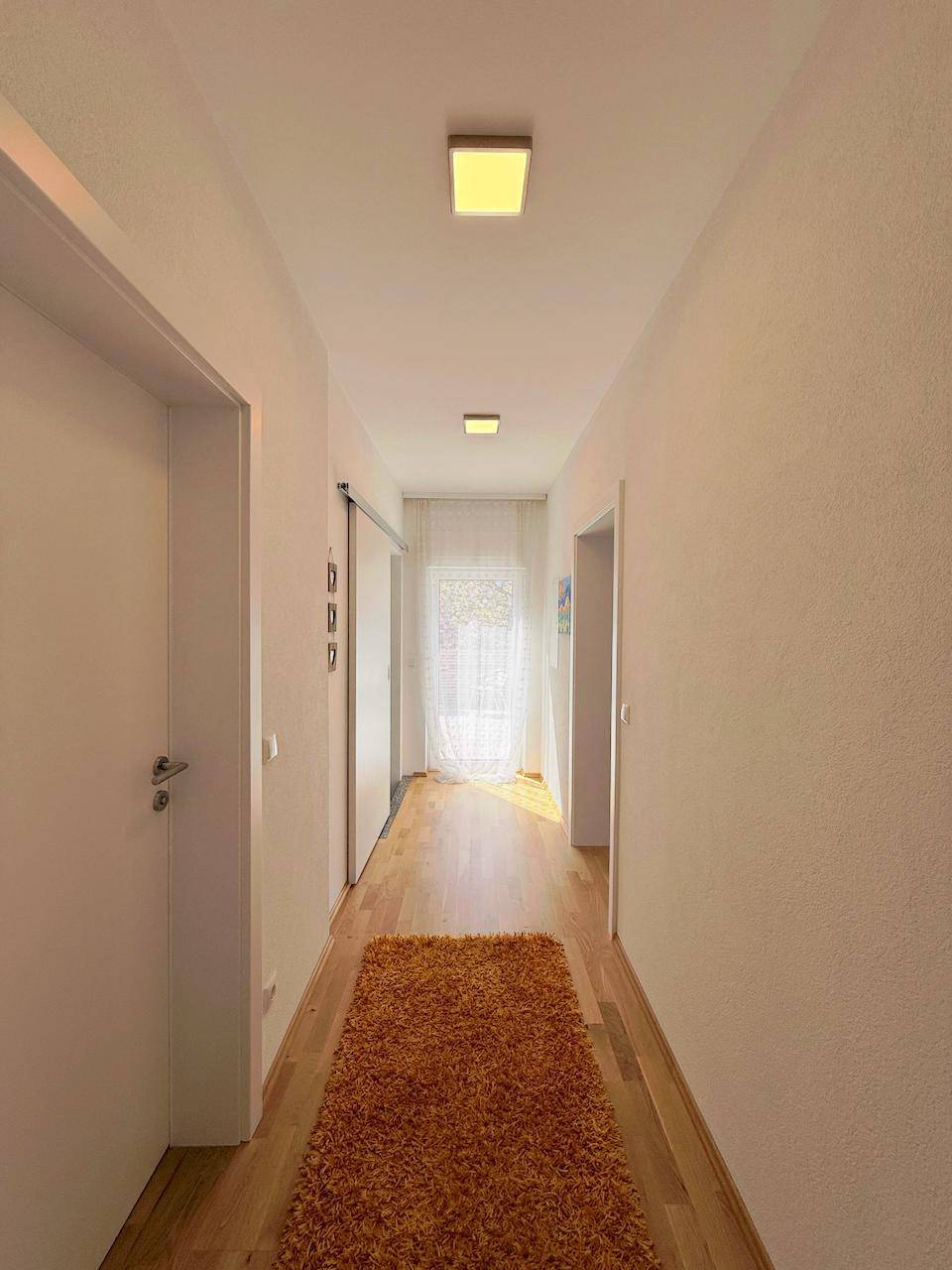 Heller Flur mit Parkettboden und modernen Deckenleuchten, der zu einem Fenster führt.
