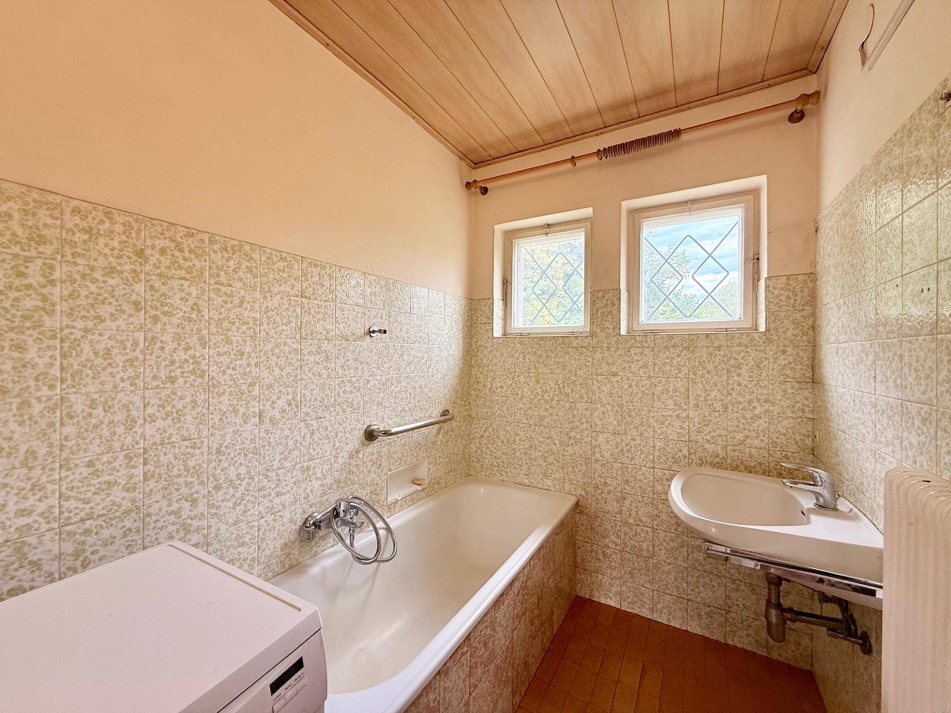 Badezimmer mit Badewanne, Waschbecken und gefliesten Wänden, bietet zwei kleine Fenster.