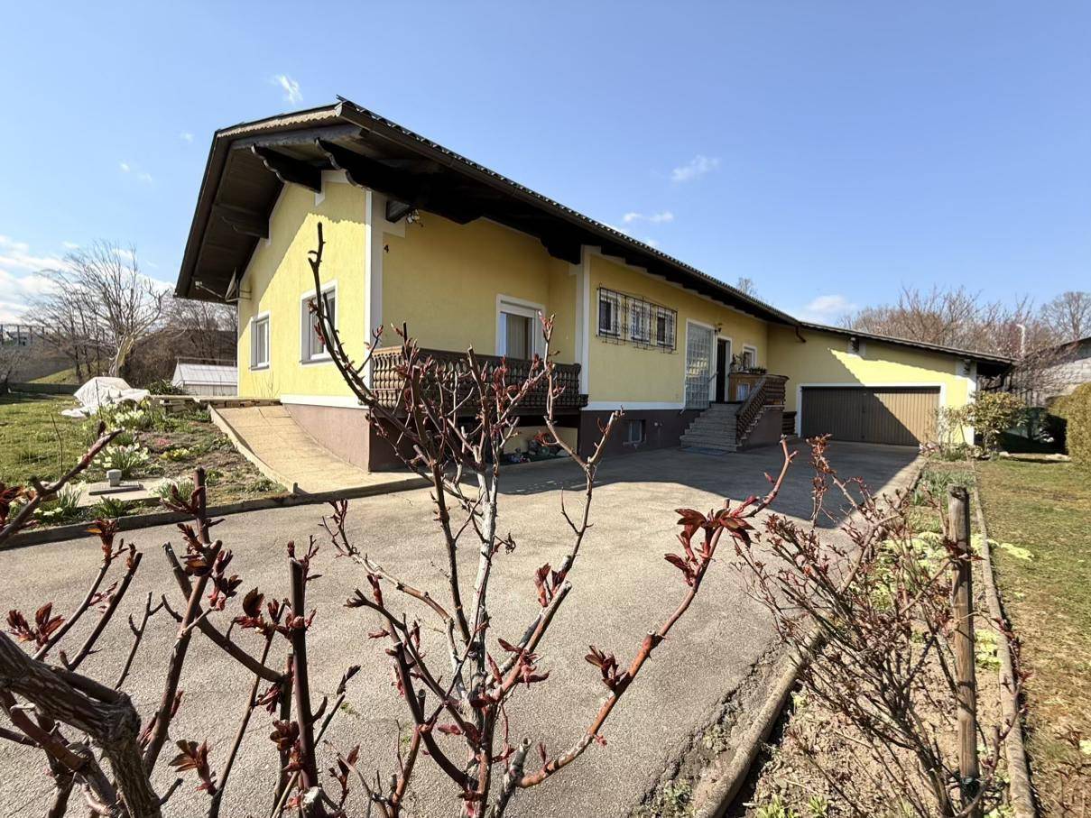 Einfahrt zum gelben Haus mit einem kleinen Balkon und umliegenden Pflanzen unter blauem Himmel.