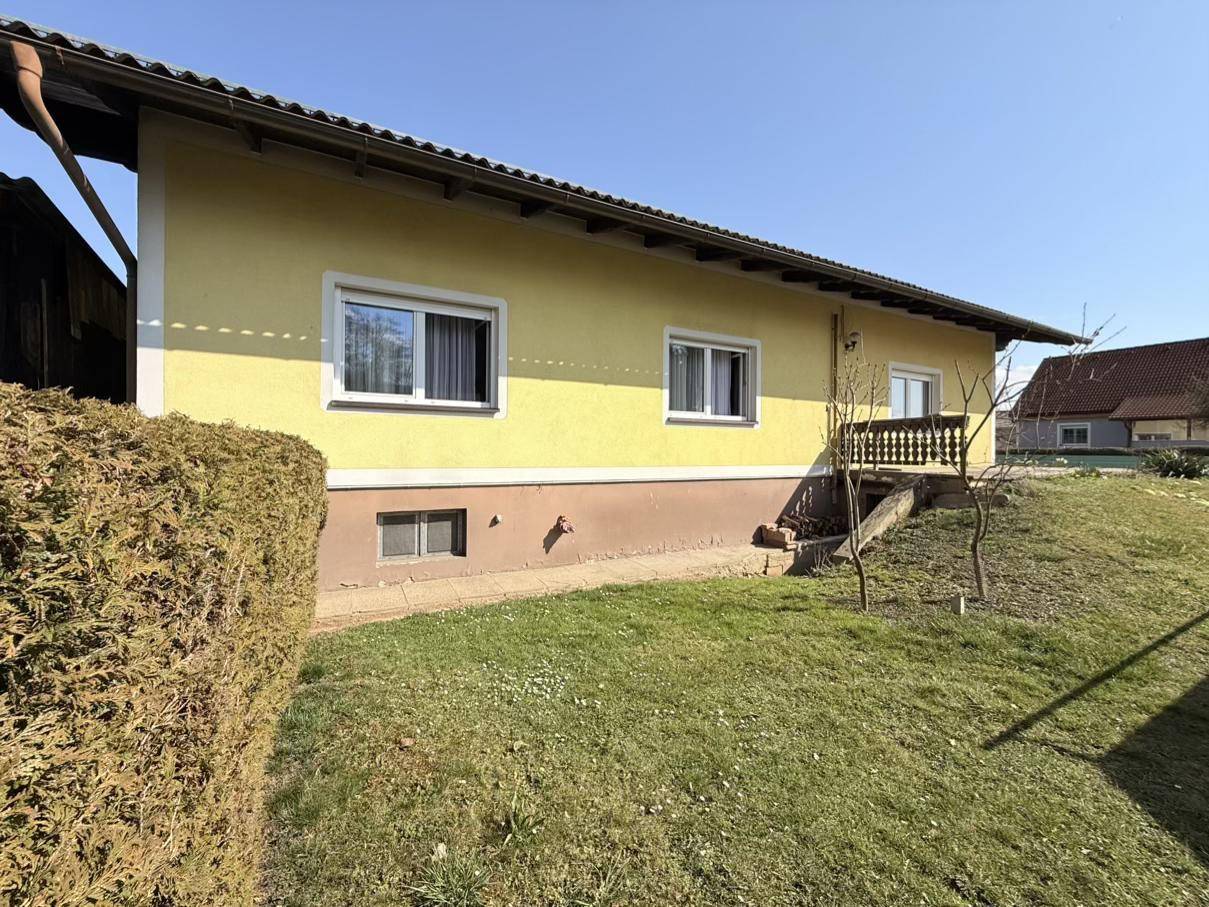 Seitenansicht des gelben Hauses mit einer gepflegten Hecke und einem grünen Rasenbereich im Sonnenschein.