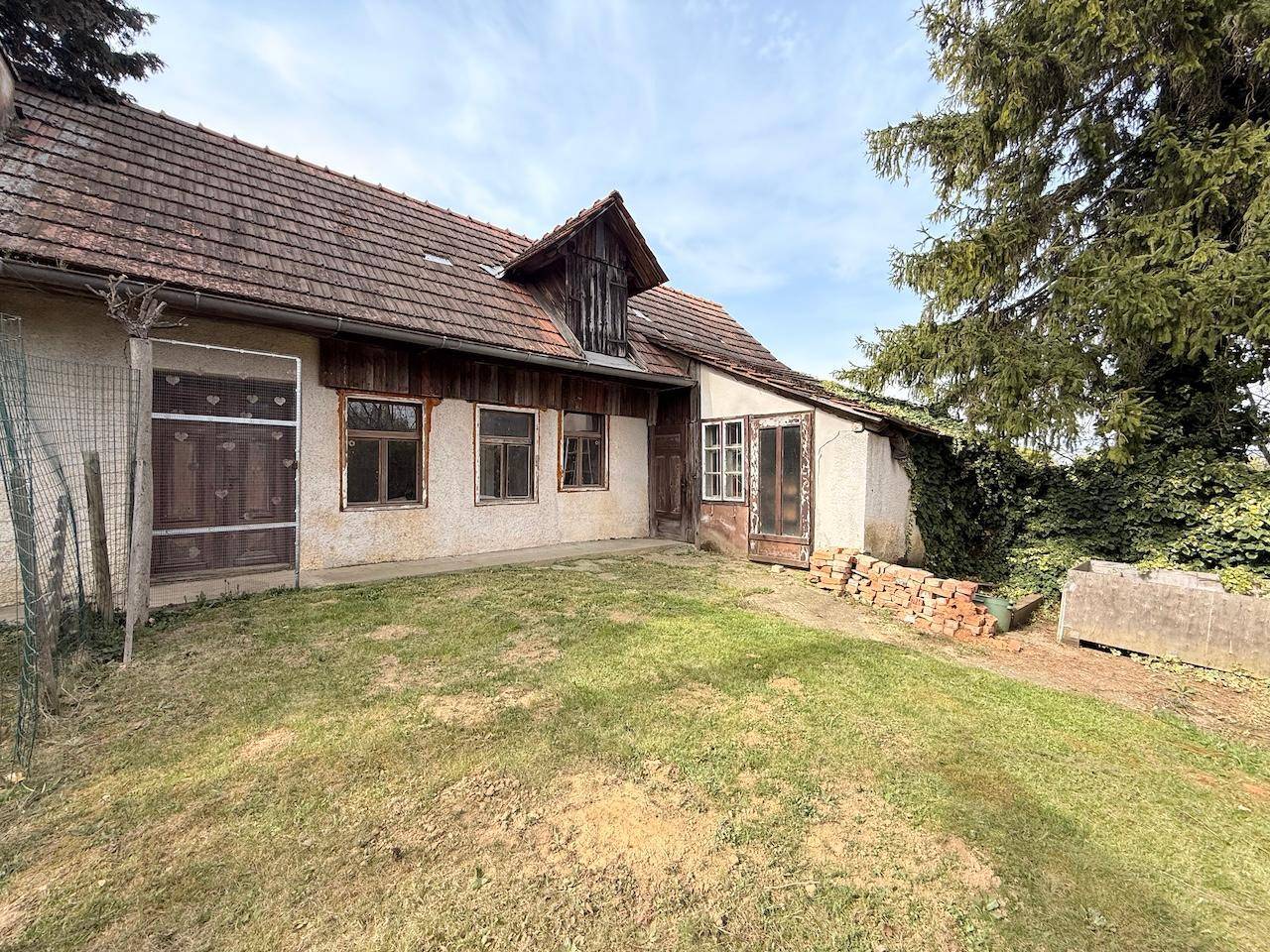 Rückseite des Hauses mit Rasenfläche und alten Fenstern, umgeben von Bäumen.