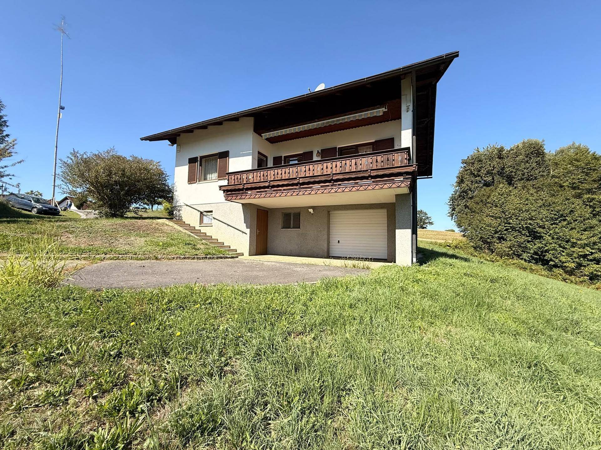 Das Haus von der Rückseite mit Balkon, Garage und Zufahrt, umgeben von grüner Natur.
