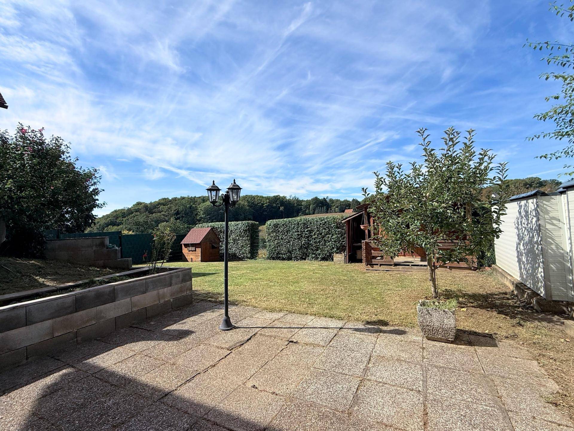 Weitläufiger Garten mit Rasen, Gartenhaus und Obstbaum unter blauem Himmel.